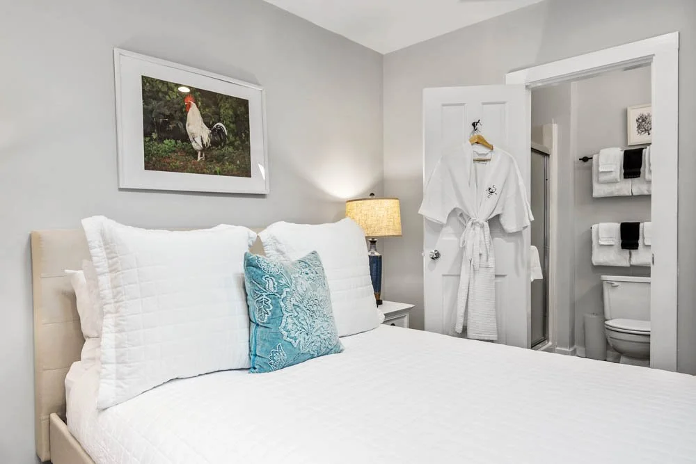 A bedroom with a white bed, turquoise pillow, framed rooster picture on the wall, and a side table with a lamp. A robe hangs on the door leading to a bathroom with shelves of towels and a toilet.