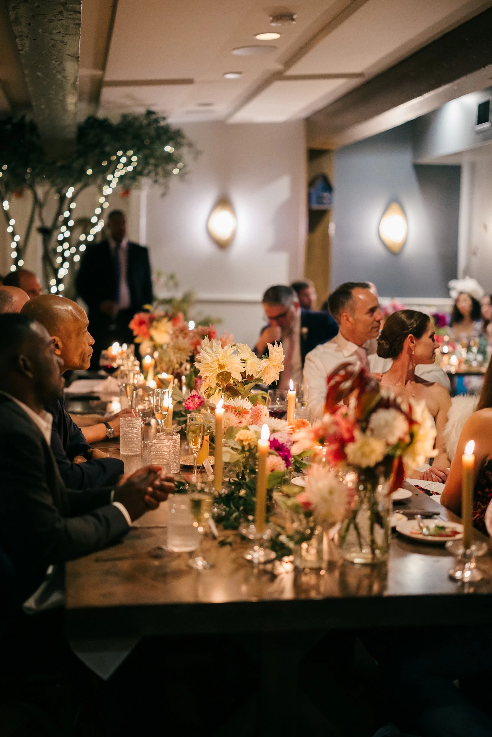 People attending a wedding reception at Lambert's Cove Inn on Martha's Vineyard with floral centerpieces and lit candles, sitting at a long table.