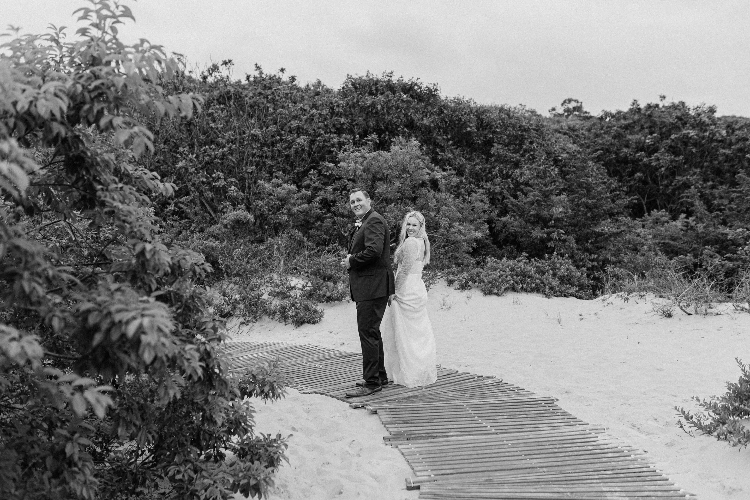 A black-and-white photo of a bride and groom on a sandy beach, standing on a wooden walkway, with dense bushes and trees in the background. The groom is in a suit, and the bride is in a wedding dress, both smiling.