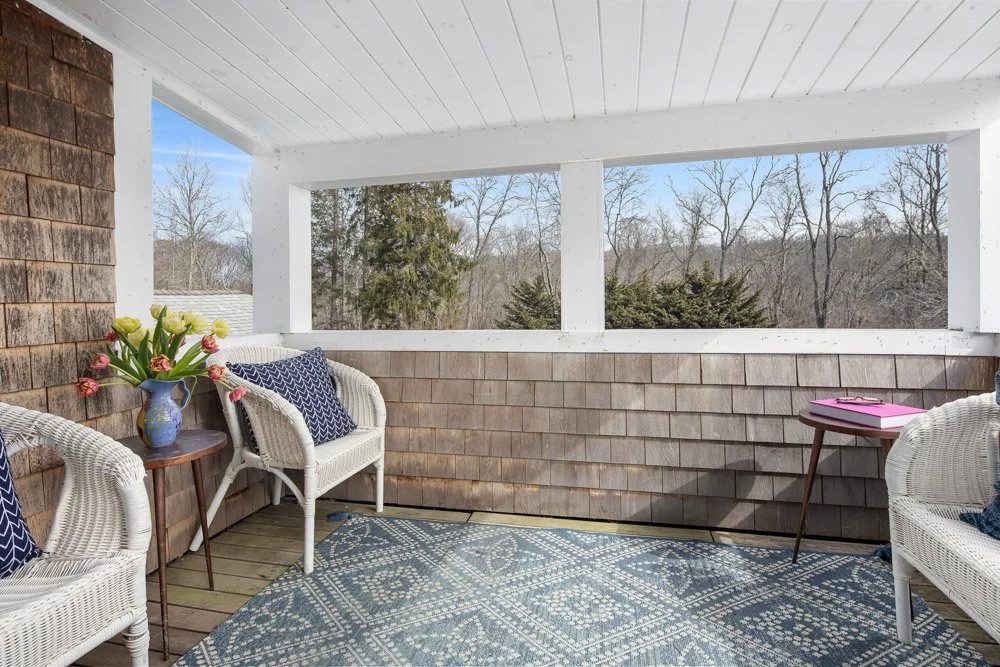 Sun porch with wicker chairs, a blue rug, and a view of leafless trees outside.