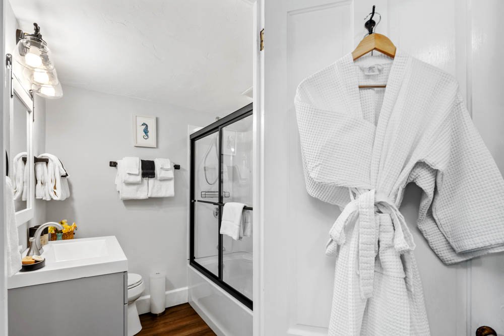 Bathroom at Lambert's Cove Inn on Martha's Vineyard with white walls, a vanity with a sink, a toilet, and a shower with a glass door. Towels are hanging on hooks, and a white bathrobe is hanging on a door. The inn hosts indoor and outdoor weddings. 