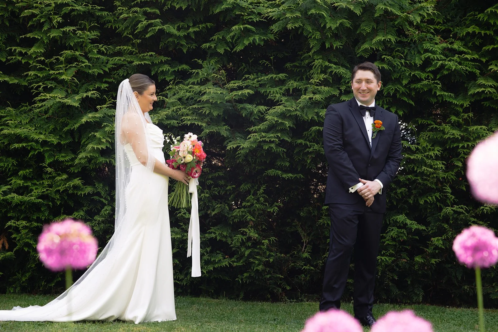 A bride and groom stand outdoors in front of green foliage, exchanging vows or sharing a moment during their wedding ceremony. The bride in a white gown holds a bouquet of pink, white, and red flowers, and the groom in a dark tuxedo smiles.