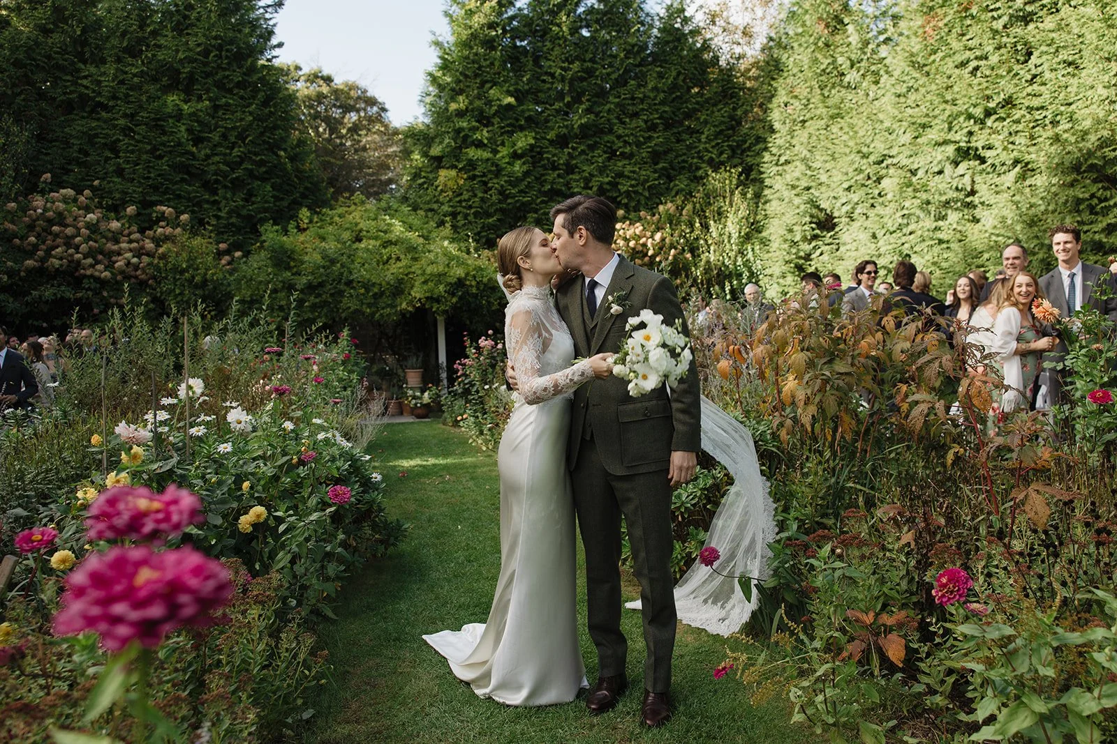 A bride and groom kissing at their wedding ceremony in a lush garden surrounded by colorful flowers and guests in the background at Lambert's Cove Inn on Martha's Vineyard.