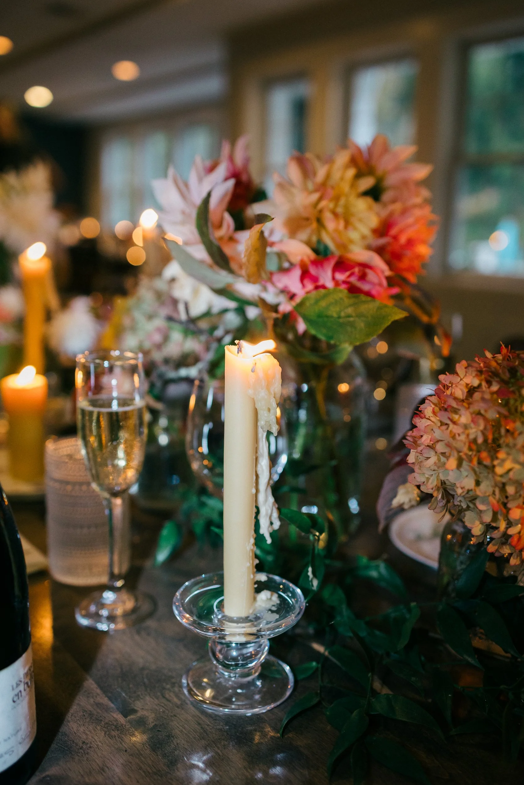A table decorated with pink and orange flowers, lit candles, and drinks, set in a cozy indoor setting with large windows at Lambert's Cove Inn on Martha's Vineyard.