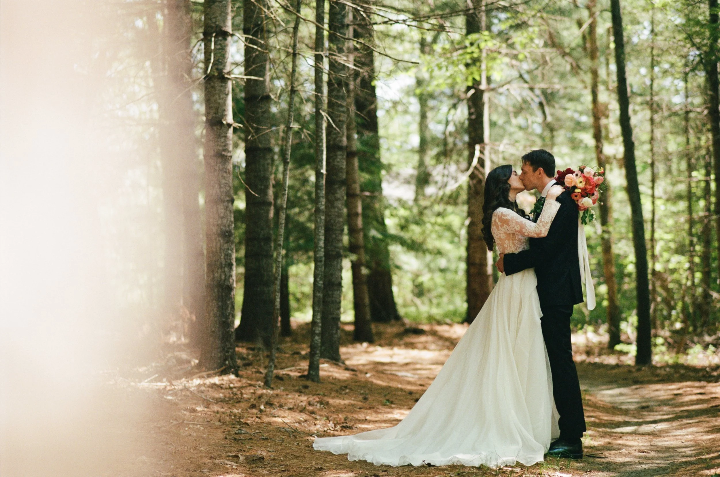 A bride and groom share a kiss in a forest during their wedding at Lambert's Cove Inn on Martha's Vineyard, with the bride wearing a white lace wedding dress holding a bouquet of flowers.