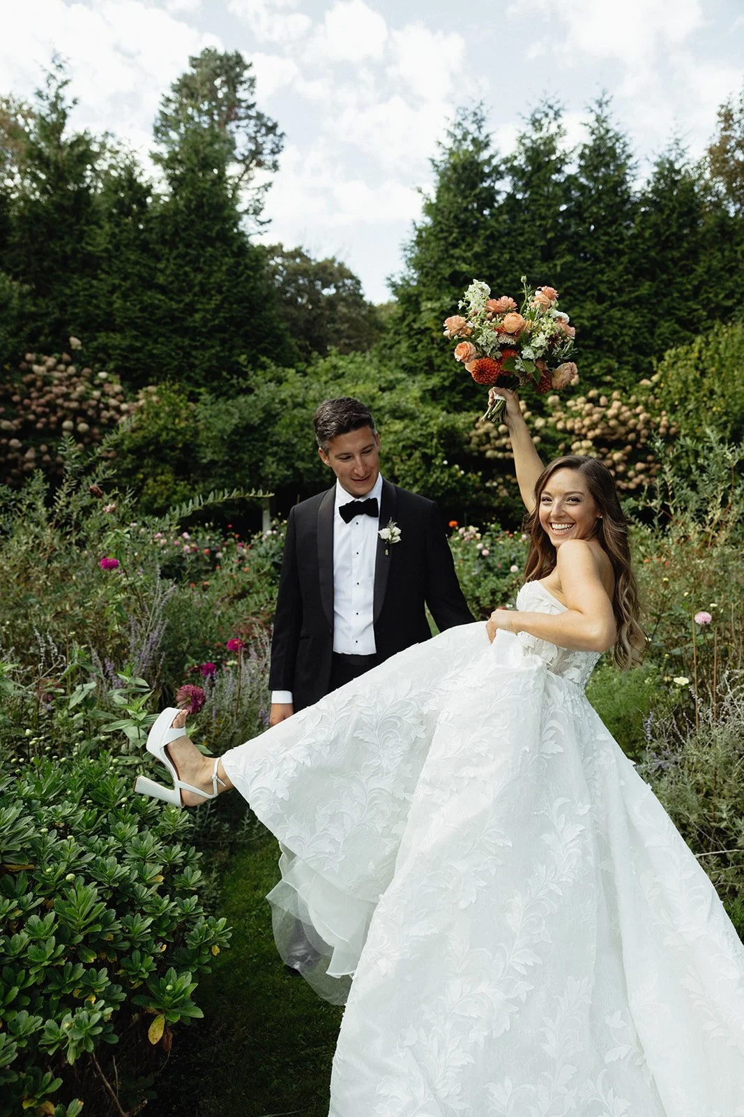 A bride and groom celebrating outdoors during a wedding at Lambert's Cove Inn on Martha's Vineyard, with the bride lifting her white dress and holding a large bouquet, smiling, in the English garden with green trees and colorful flowers.