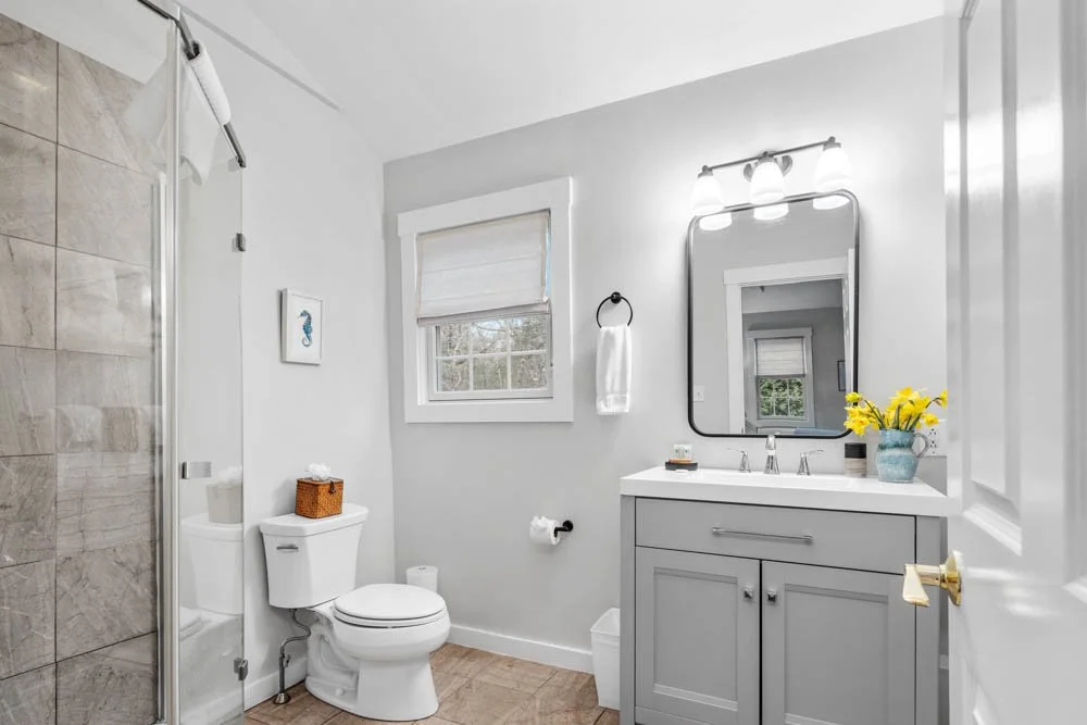 A clean, bright bathroom featuring a glass-enclosed shower, a toilet with a tissue box on top, a small window with blinds, a wall-mounted towel ring with a towel, a mirror above a gray vanity with a yellow flower arrangement, and sleek lighting fixtu