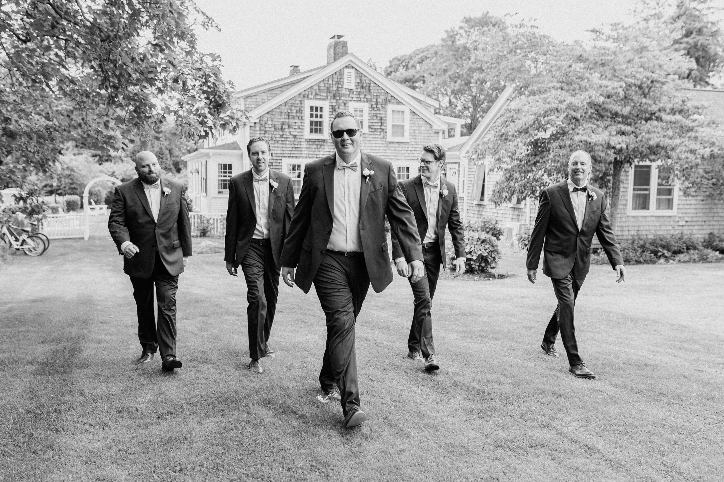 A group of five men walking on a lawn before a wedding at Lambert's Cove Inn on Martha's Vineyard, dressed in suits with ties or bow ties, in front of a house with trees and shrubs, during daytime in black and white.