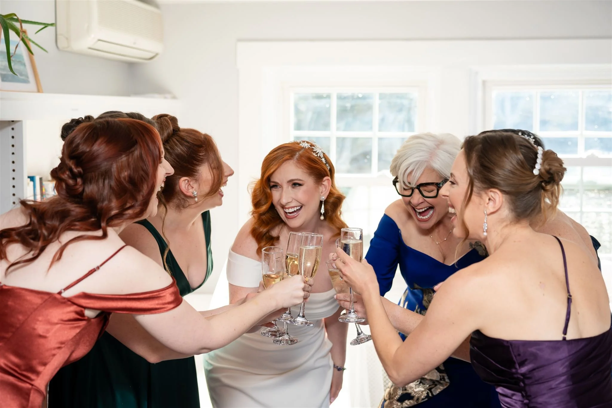 A group of women dressed in formal evening gowns celebrating together, holding champagne glasses, in a bright room with large windows at Lambert's Cove Inn on Martha's Vineyard.