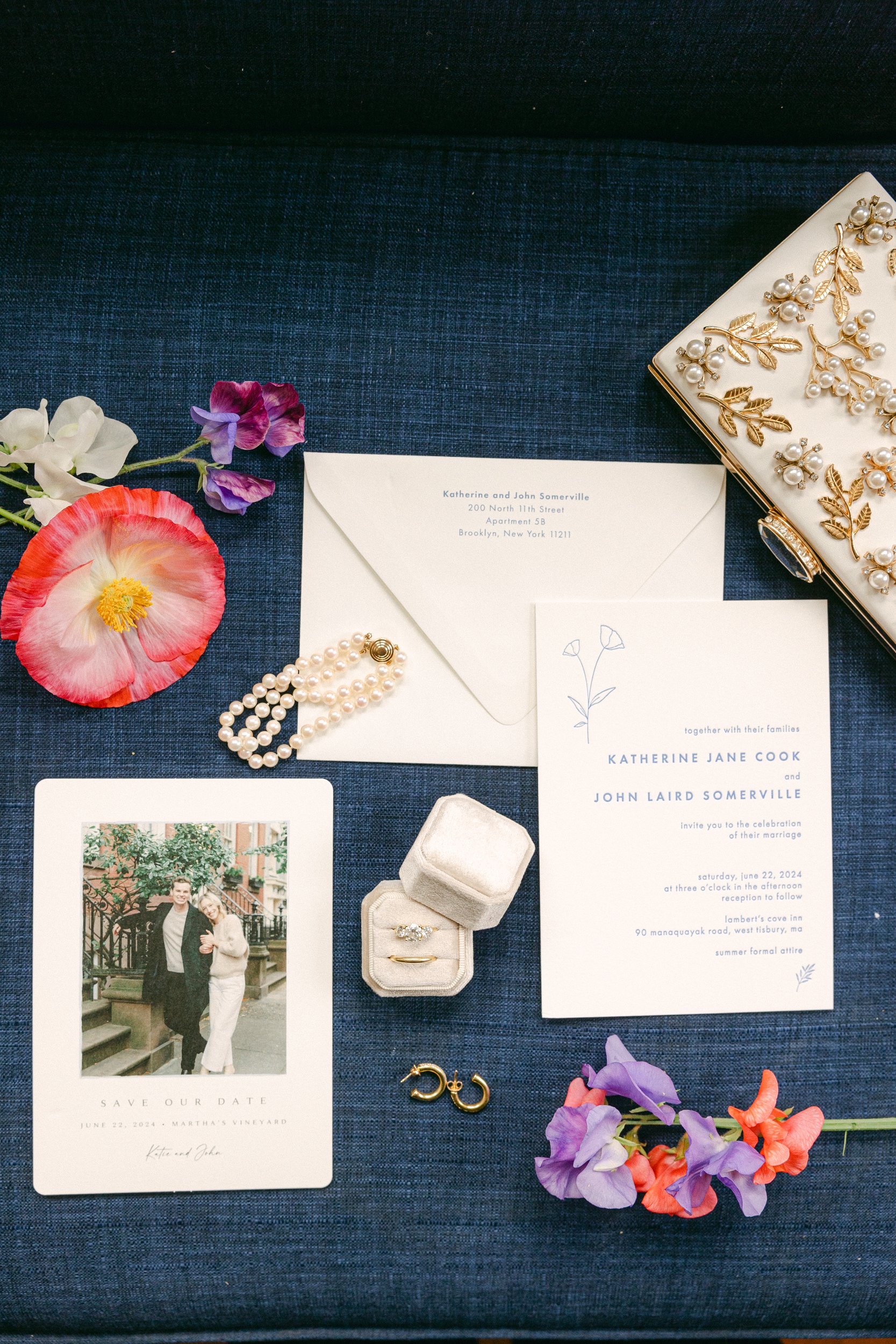 Wedding invitation suite with floral accents, jewelry, a photograph of the couple, and a ring box on a dark blue fabric background.