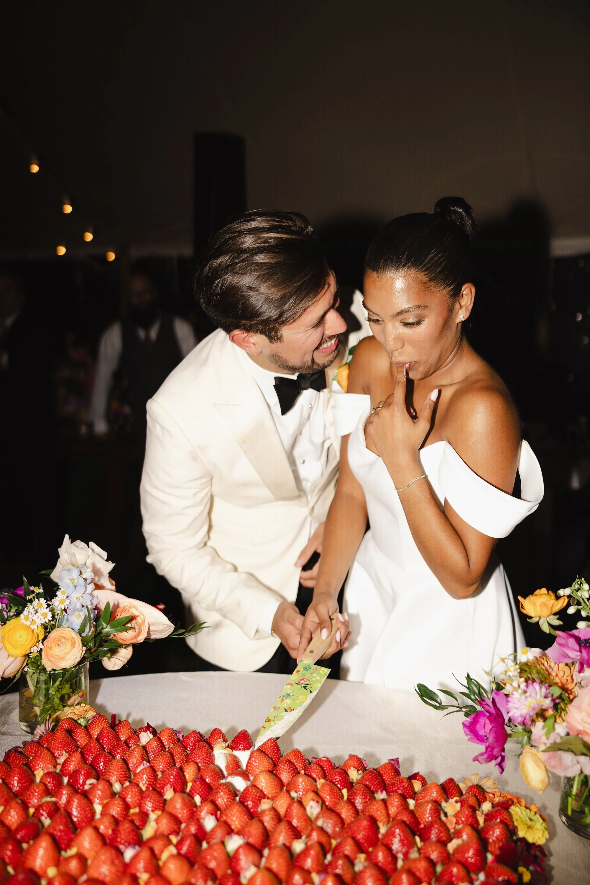 A newlywed couple cutting their wedding cake at Lambert's Cove Inn on Martha's Vineyard surrounded by fresh strawberries and colorful flowers.