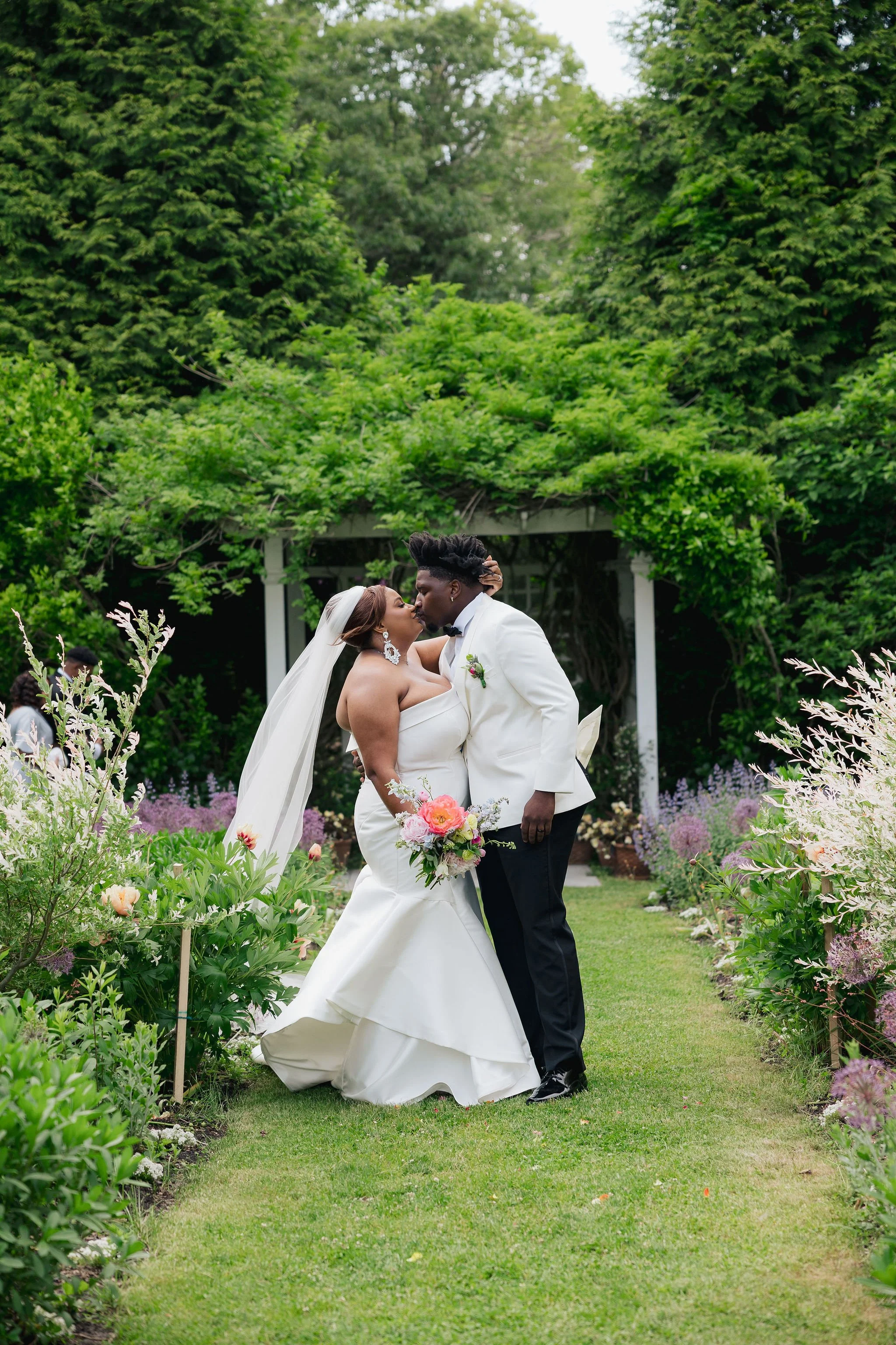 A bride and groom kissing in a lush garden during their wedding ceremony, with greenery and flowers surrounding them at Lambert's Cove Inn on Martha's Vineyard.
