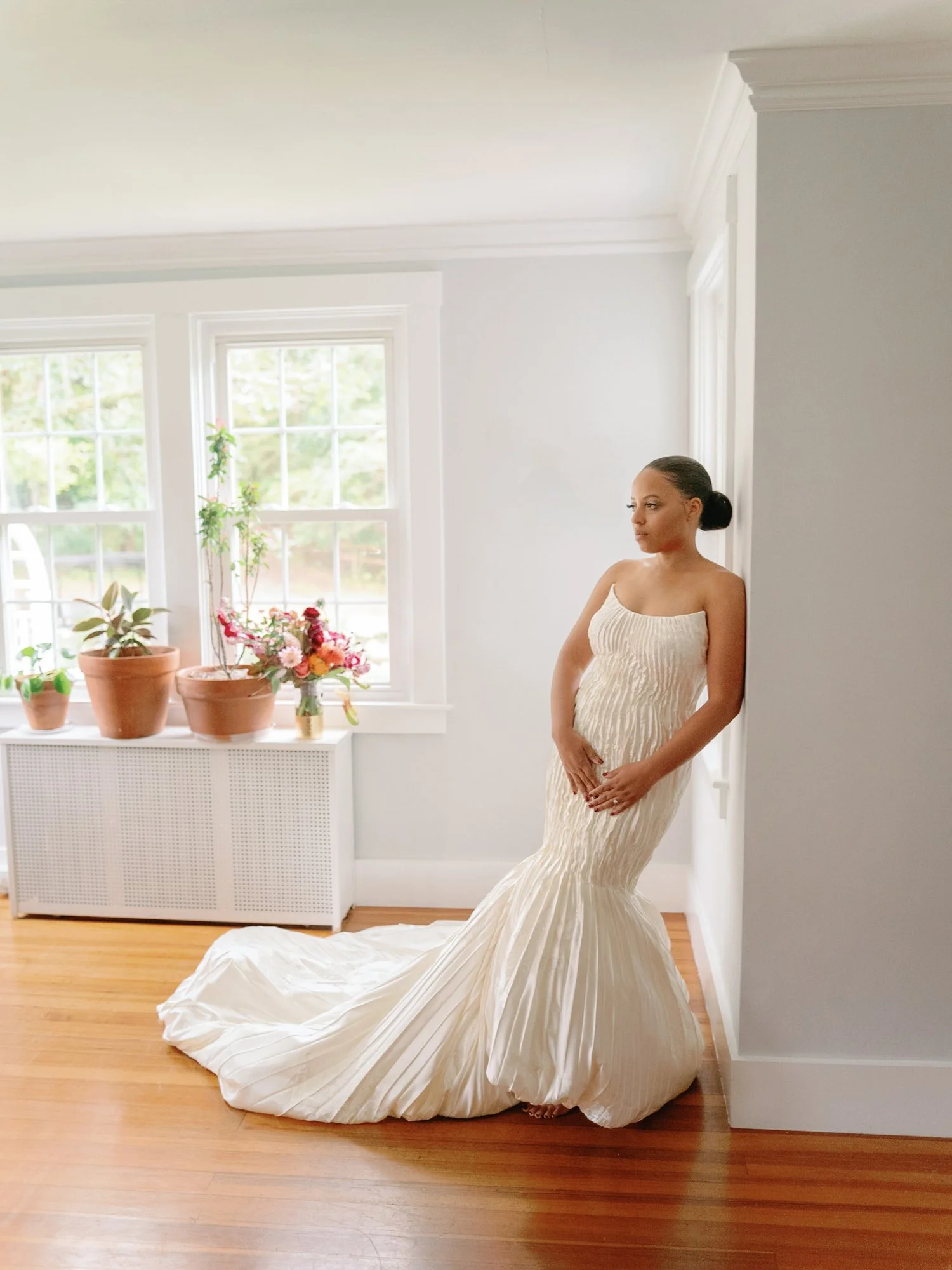 A woman in a cream-colored, pleated, strapless gown stands barefoot in a bright, white room with wooden floors, leaning against a wall near two large windows with plants and flowers on a white radiator cover.