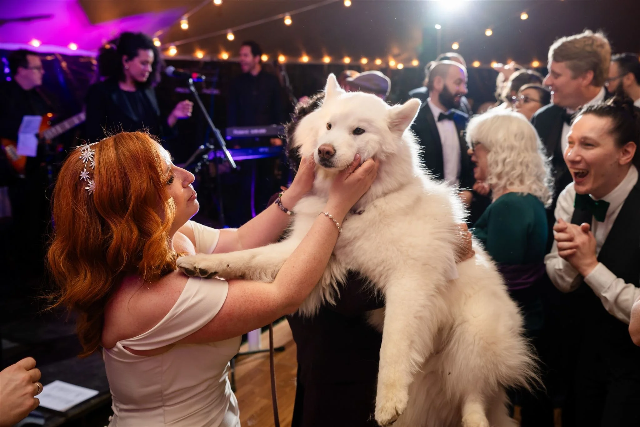 A woman with red hair and a floral headband holding a large white dog at wedding reception at Lambert's Cove Inn on Martha's Vineyard with other people, some dressed formally, dancing and enjoying live music under string lights.