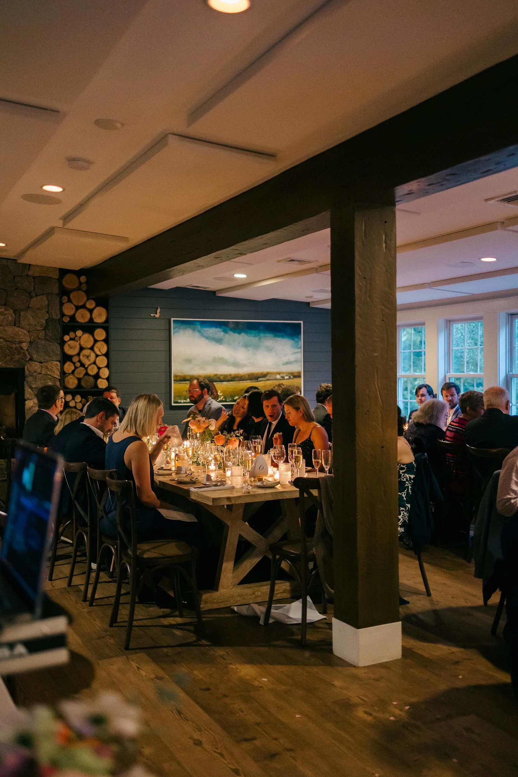 People gathered at wedding reception at Lambert's Cove Inn on Martha's Vineyard in a well-lit room with natural light coming from large windows, decorated with flowers and artwork on the walls.
