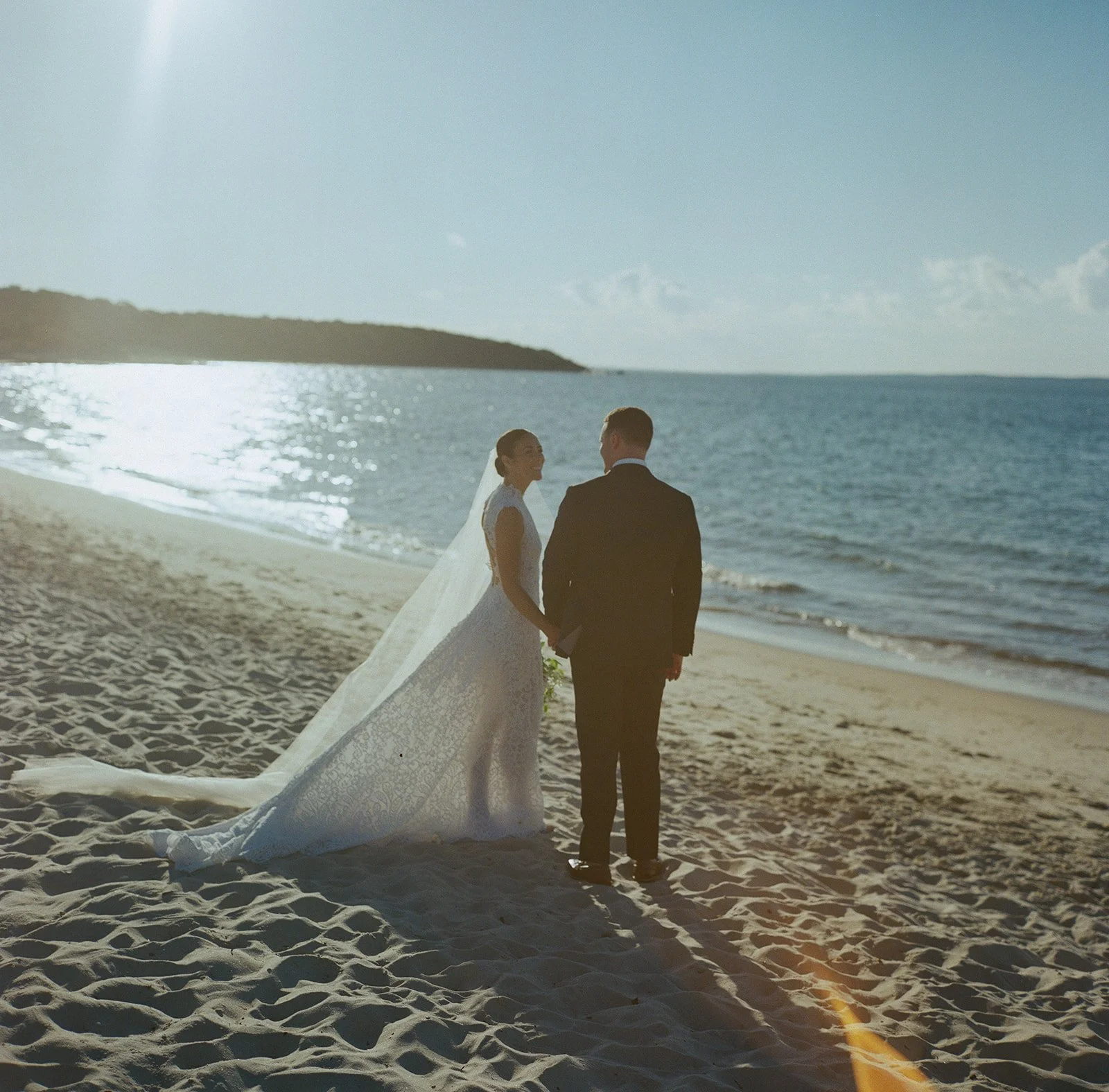 A bride and groom standing hand in hand on a sandy beach with the ocean and distant land in the background during sunset or sunrise during a wedding at Lambert's Cove Inn on Martha's Vineyard.