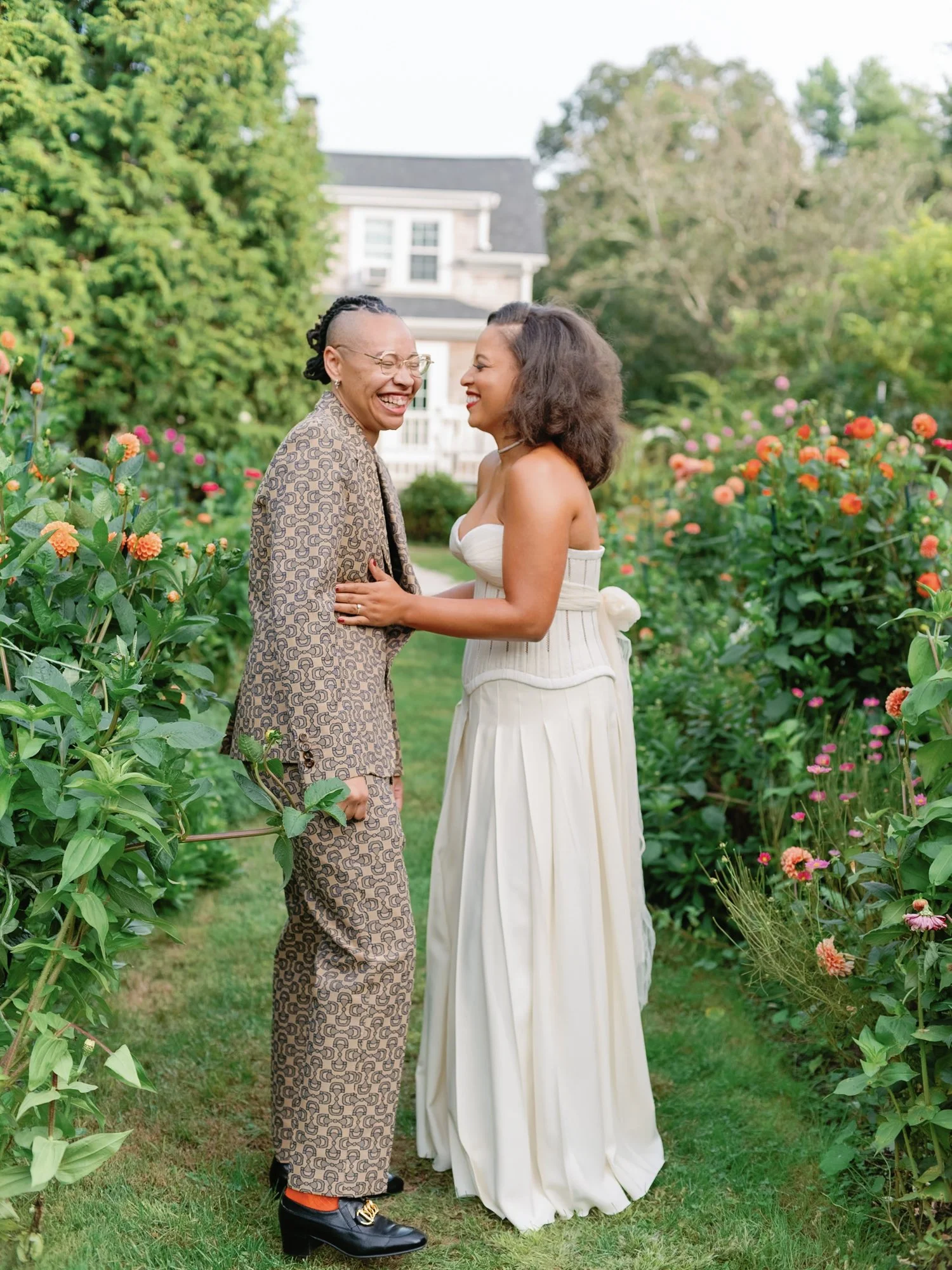 Two women are standing face to face, smiling and laughing together in a garden with colorful flowers. One woman has short hair with a shaved side, wearing glasses, a patterned suit, and black boots. The other woman has shoulder-length curly hair and 