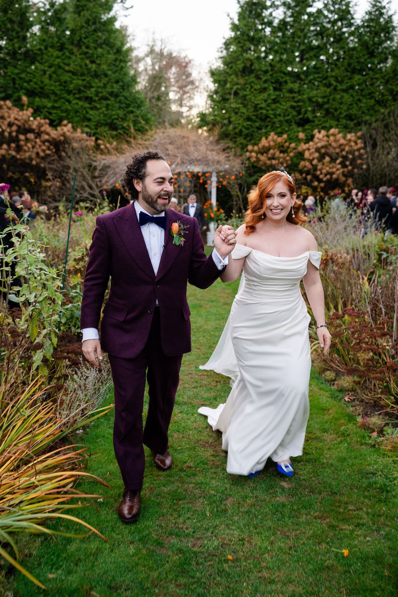 A newlywed couple walking outdoors during a reception at Lambert's Cove Inn on Martha's Vineyard, smiling and holding hands with a garden and other guests in the background.