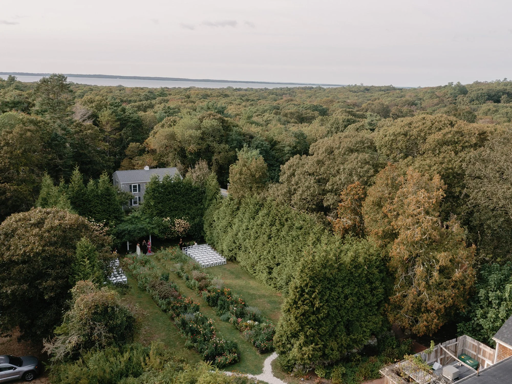 Aerial view of the lush English garden at Lambert's Cove Inn on Martha's Vineyard with a small house and garden in the foreground, overlooking a distant body of water under a cloudy sky.