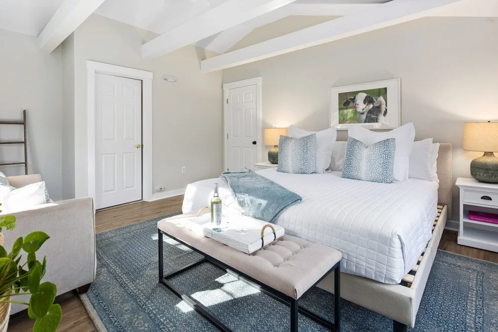 Bright bedroom with white walls and ceiling, featuring a large bed with white bedding and blue pillows, two nightstands with lamps, a bench at the foot of the bed with a tray of drinks, and artwork of a cow above the bed.