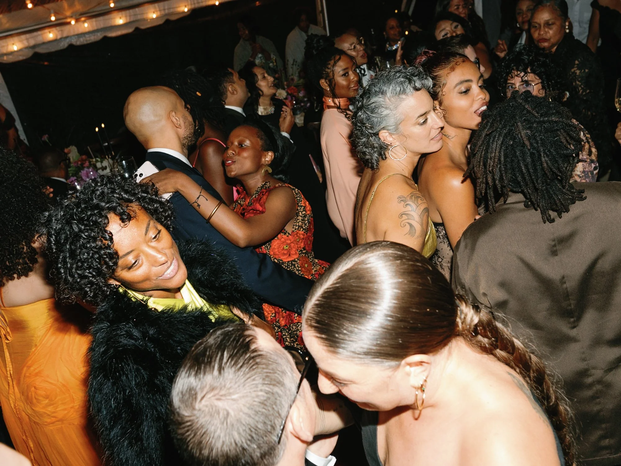 A crowded gathering of diverse people at lively wedding reception at Lambert's Cove Inn on Martha's Vineyard, closely packed together, some with tattoos and jewelry, in a well-lit setting with decorations.