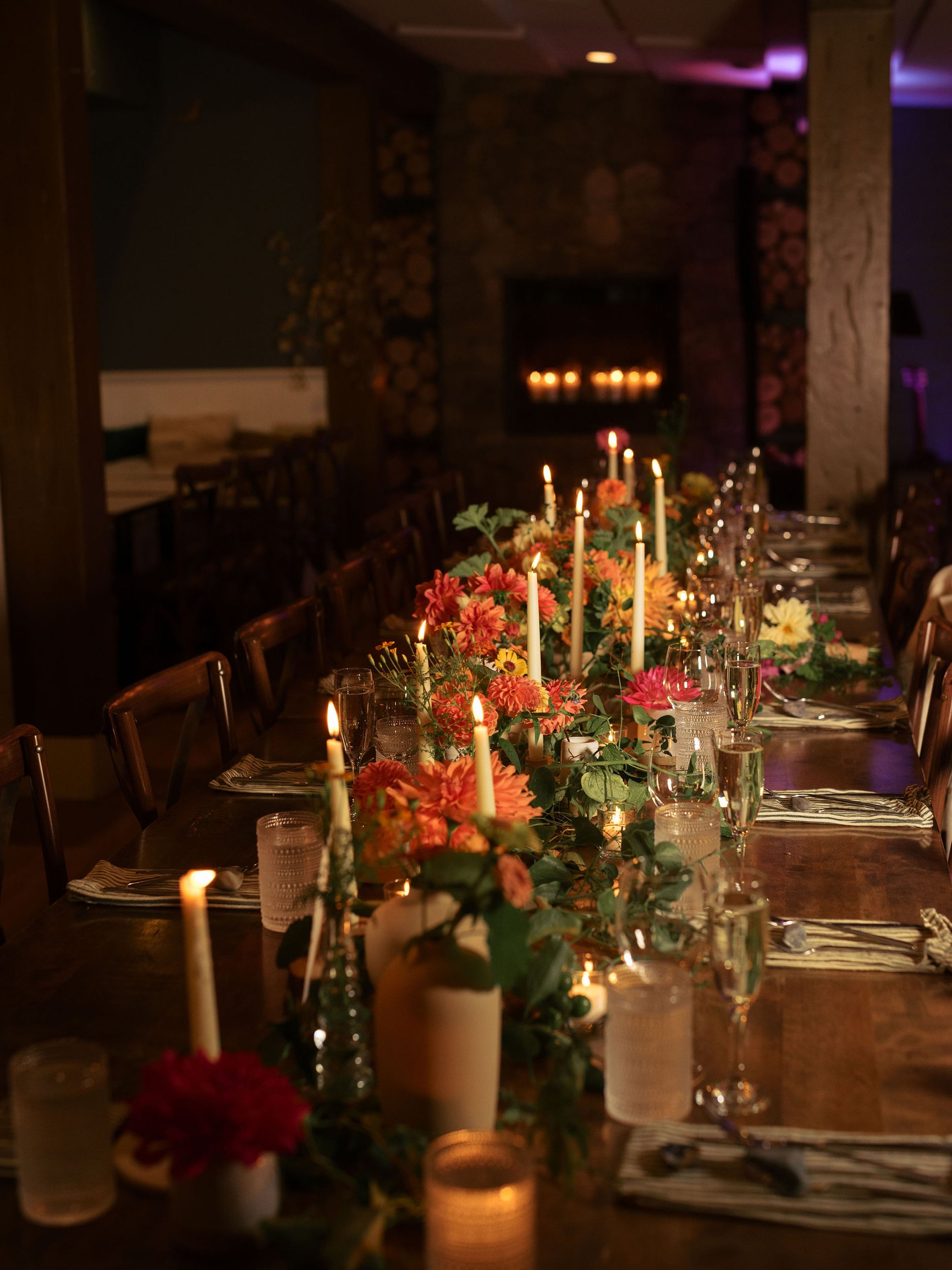 A long dining table decorated with colorful flowers and lit candles in glass holders, set for a formal dinner in a dimly lit room with a stone fireplace at Lambert's Cove Inn on Martha's Vineyard.