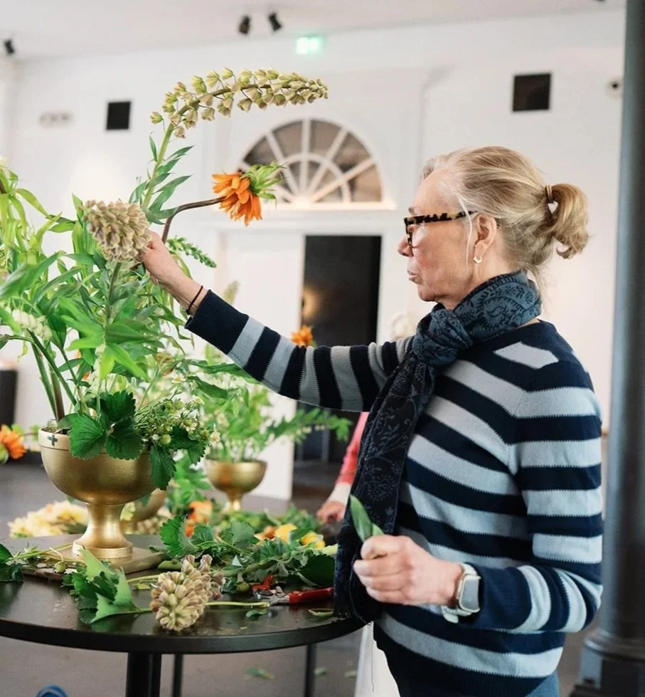 An elderly woman with blonde hair tied back, wearing glasses, a striped sweater, and a scarf, arranging flowers in a gold vase at a table.