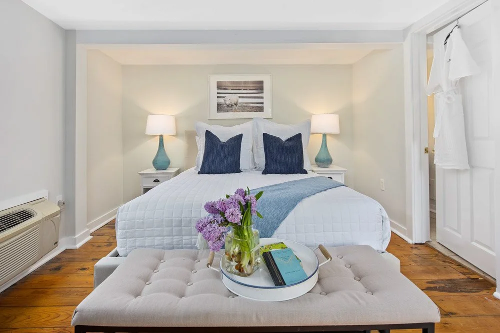 Decorated bedroom with a white bed, blue pillows, two matching lamps on side tables, a framed picture above the bed, a beige tufted bench with a tray holding purple flowers and books, hardwood floor, and a white door with a white robe hanging on it.