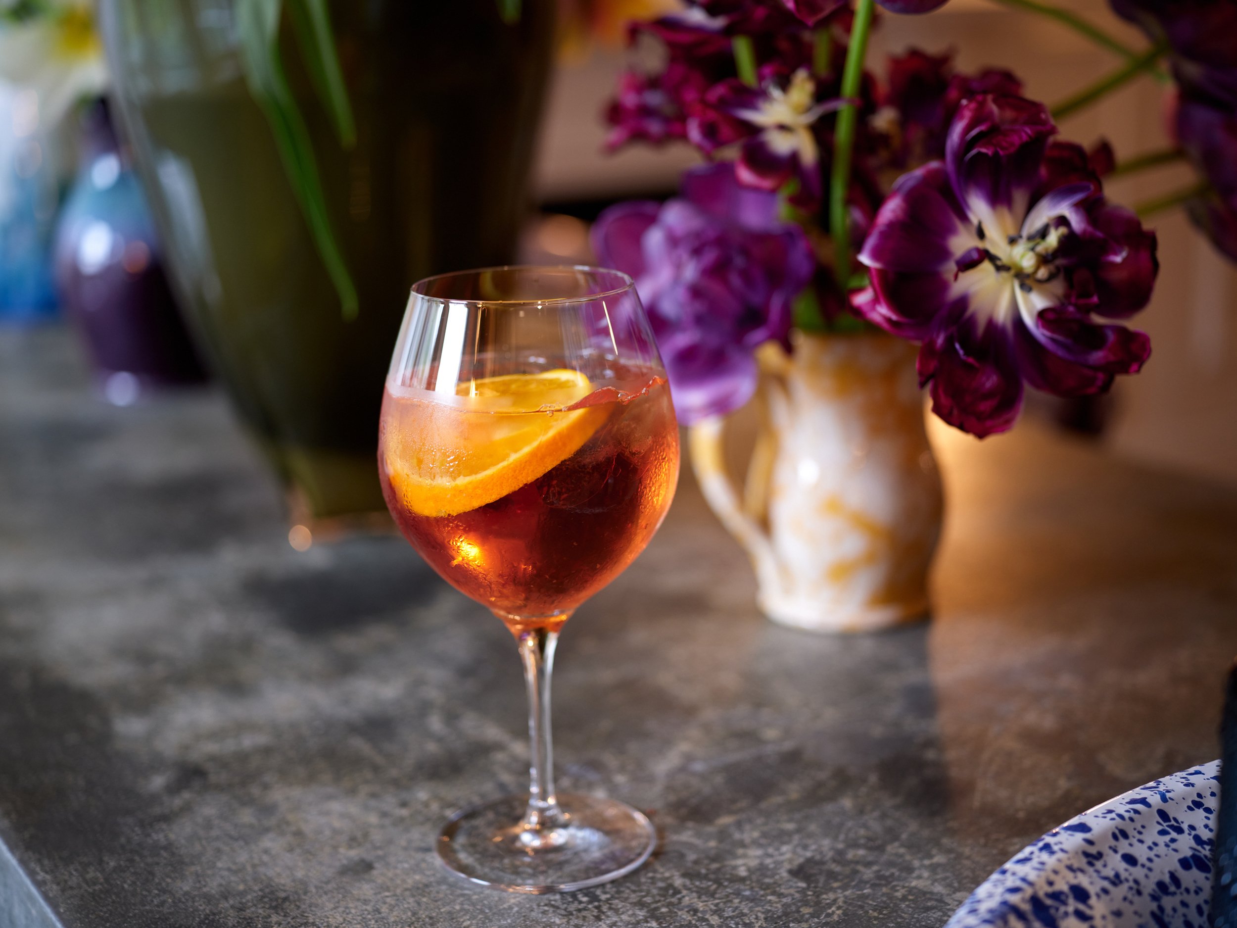 A wine glass filled with an orange-red drink garnished with an orange slice, set on a dark table with purple and pink flowers in a vase in the background.