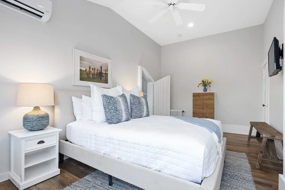 A modern bedroom with a white bed, blue and white pillows, a bedside table with a lamp, a wall-mounted TV, a small wooden bench, a wooden dresser with a flower vase, and framed wall art. The room has light gray walls, a ceiling fan, and wood flooring