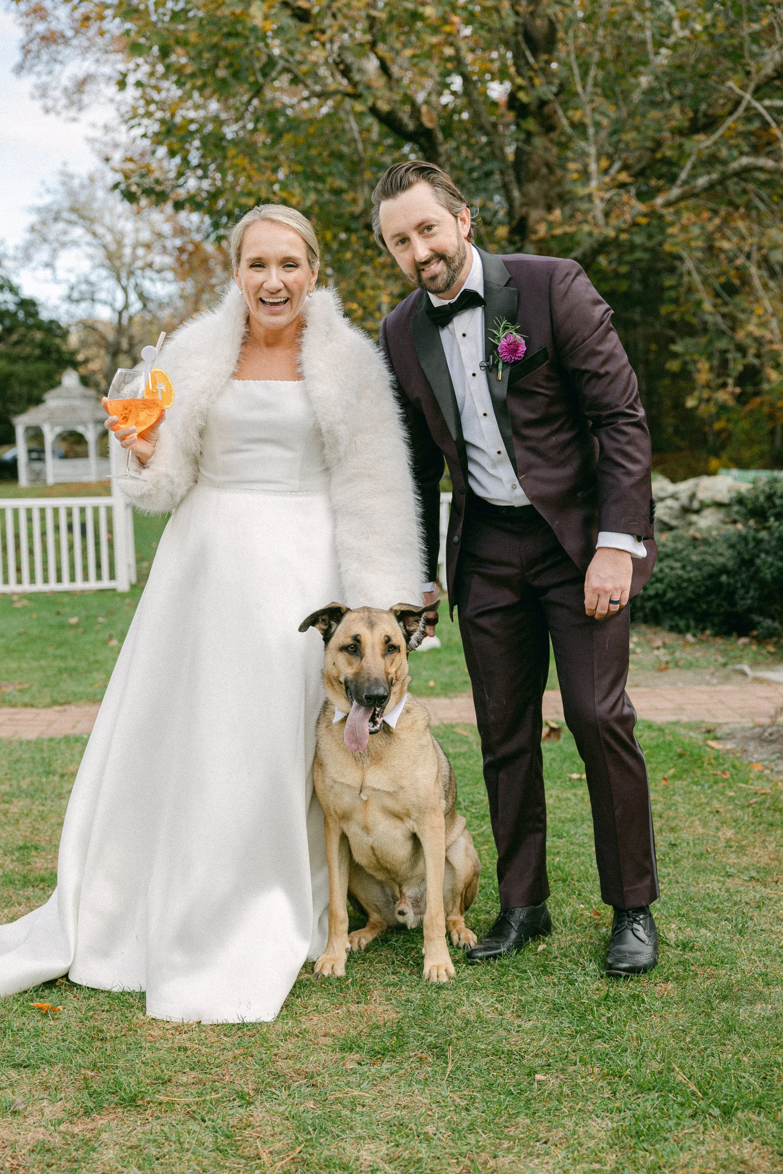 A woman in a white wedding dress and a man in a tuxedo pose with a large dog outdoors during autumn. The woman is holding a cocktail and smiling, and the man is leaning toward the dog. The background features trees with fall foliage and a white gazeb