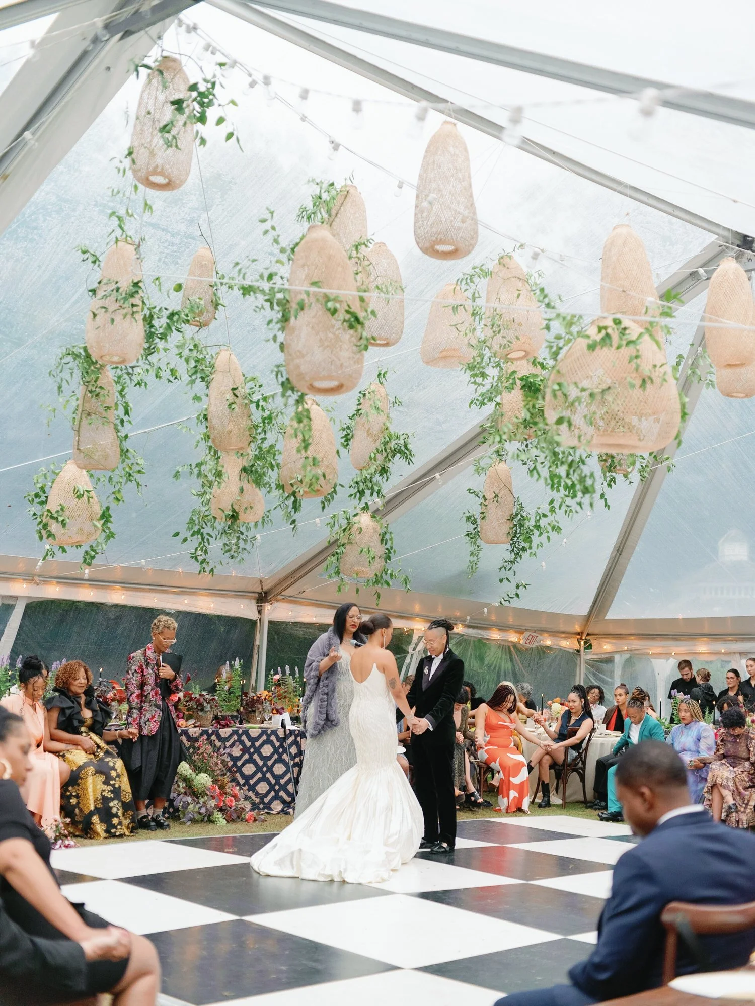 A wedding ceremony is taking place in a tent at Lambert's Cove Inn on Martha's Vineyard with decorative hanging lanterns and lights, facing a mix of seated guests and a bride and groom holding hands on a dance floor.