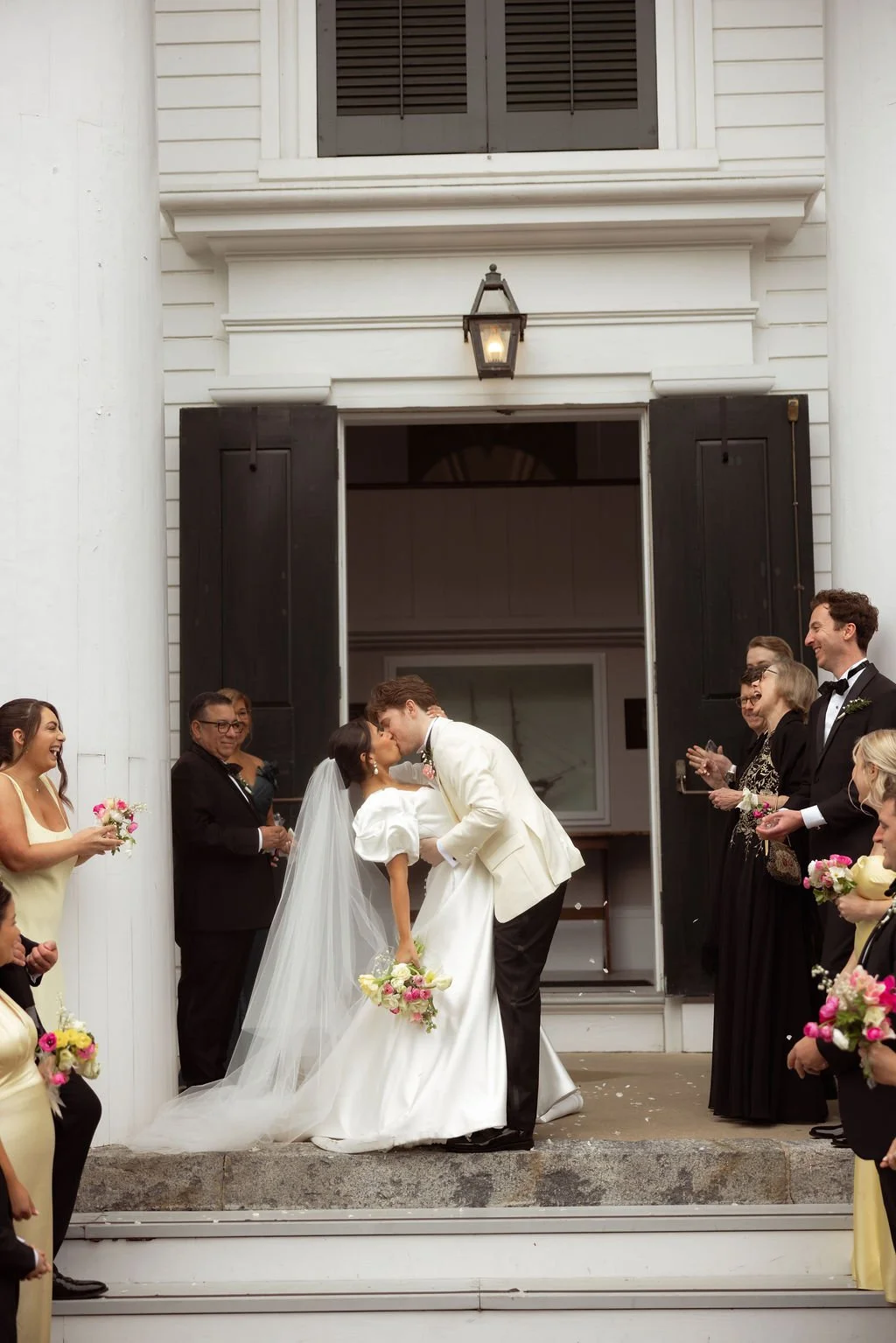 A bride and groom kiss during their wedding ceremony outside a church, surrounded by friends and family holding bouquets and applauding.
