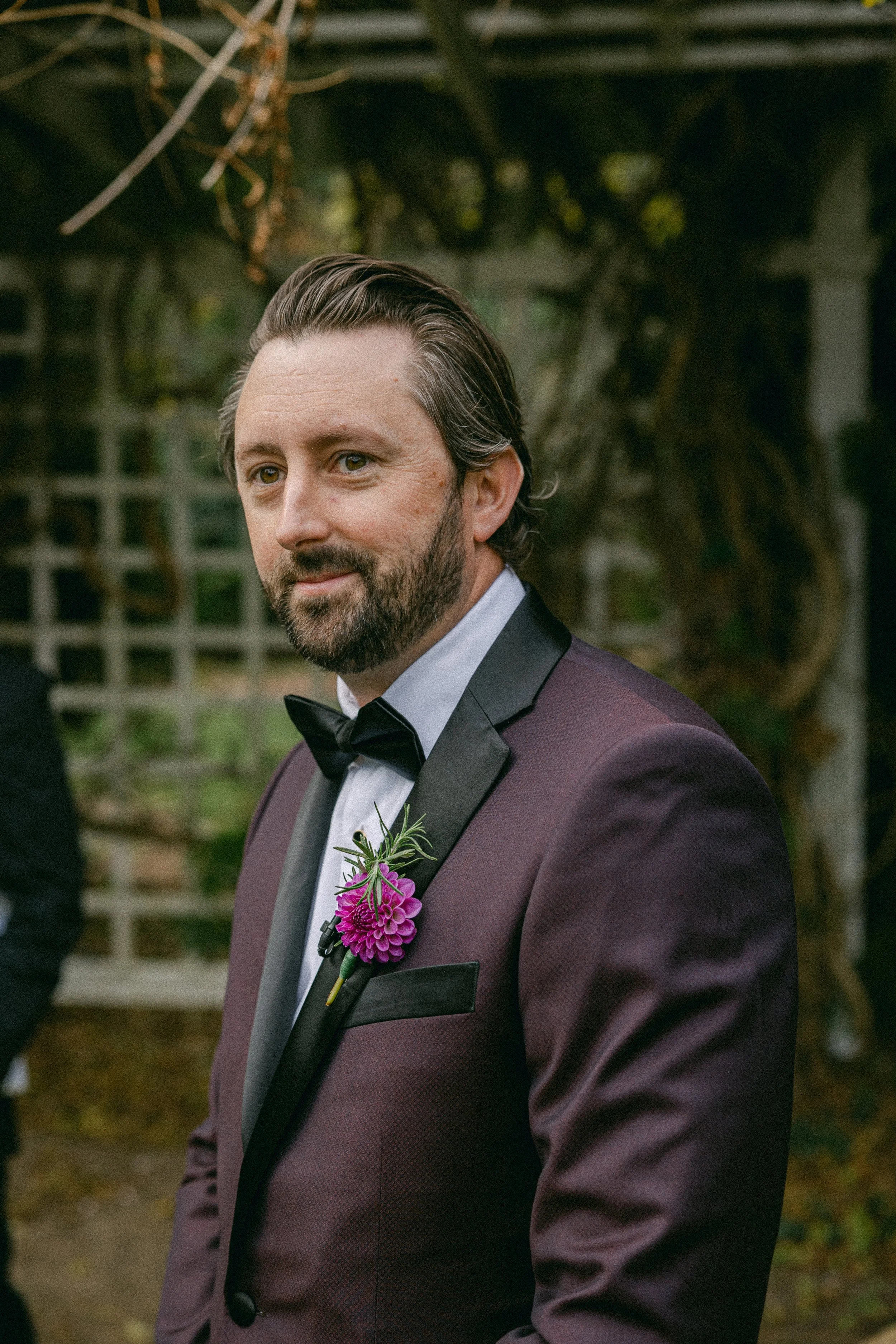 A man dressed in a tuxedo with a boutonniere stands outdoors. He has light skin, dark brown hair, and a beard. The background features greenery and a wooden lattice at Lambert's Cove Inn on Martha's Vineyard.