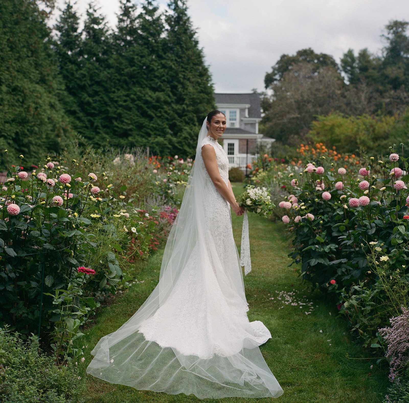 A bride in a white lace wedding dress with a long veil standing in a lush garden filled with pink and white flowers, in front of a large house and trees during a wedding reception at Lambert's Cove Inn on Martha's Vineyard.
