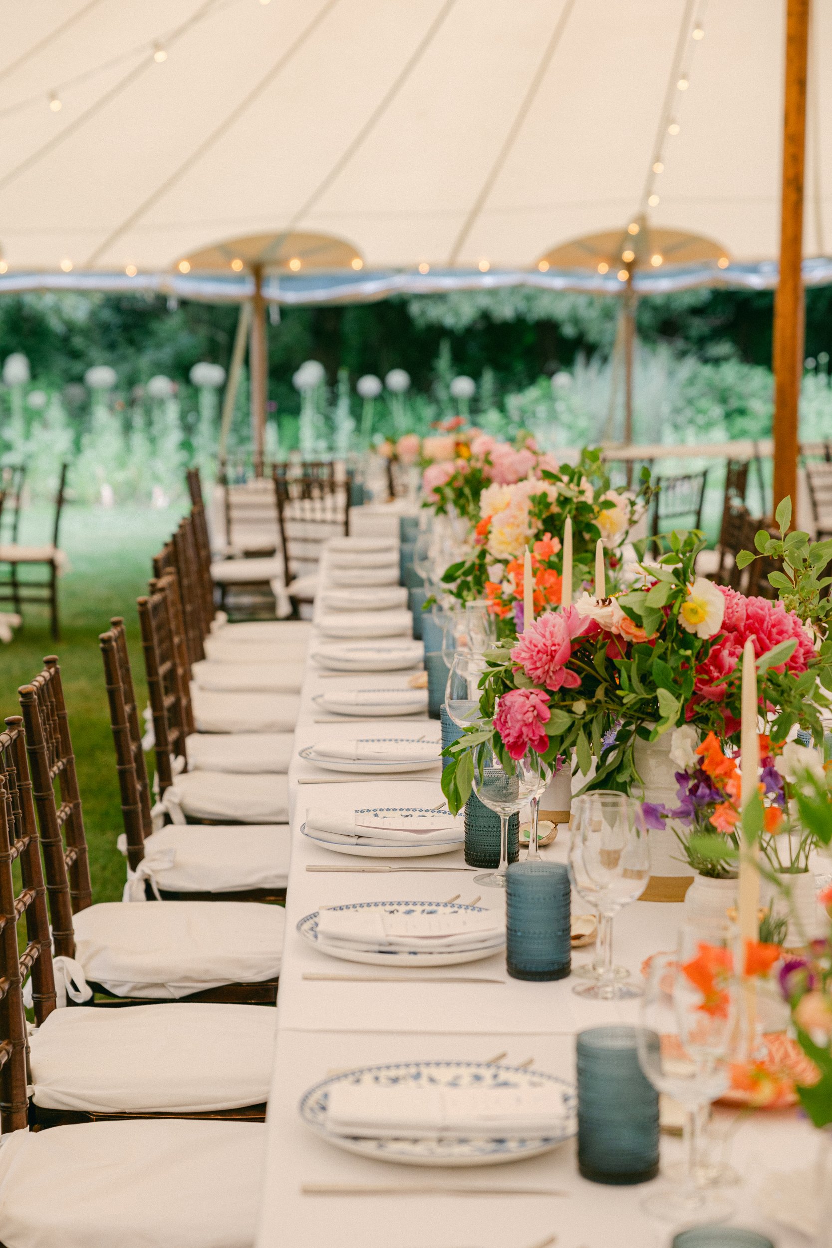 Long outdoor dining table decorated with pink, white, and orange flowers in vases, set with white plates, glasses, and napkins under a large tent with string lights, in a garden setting during a wedding at Lambert's Cove Inn on Martha's Vineyard.