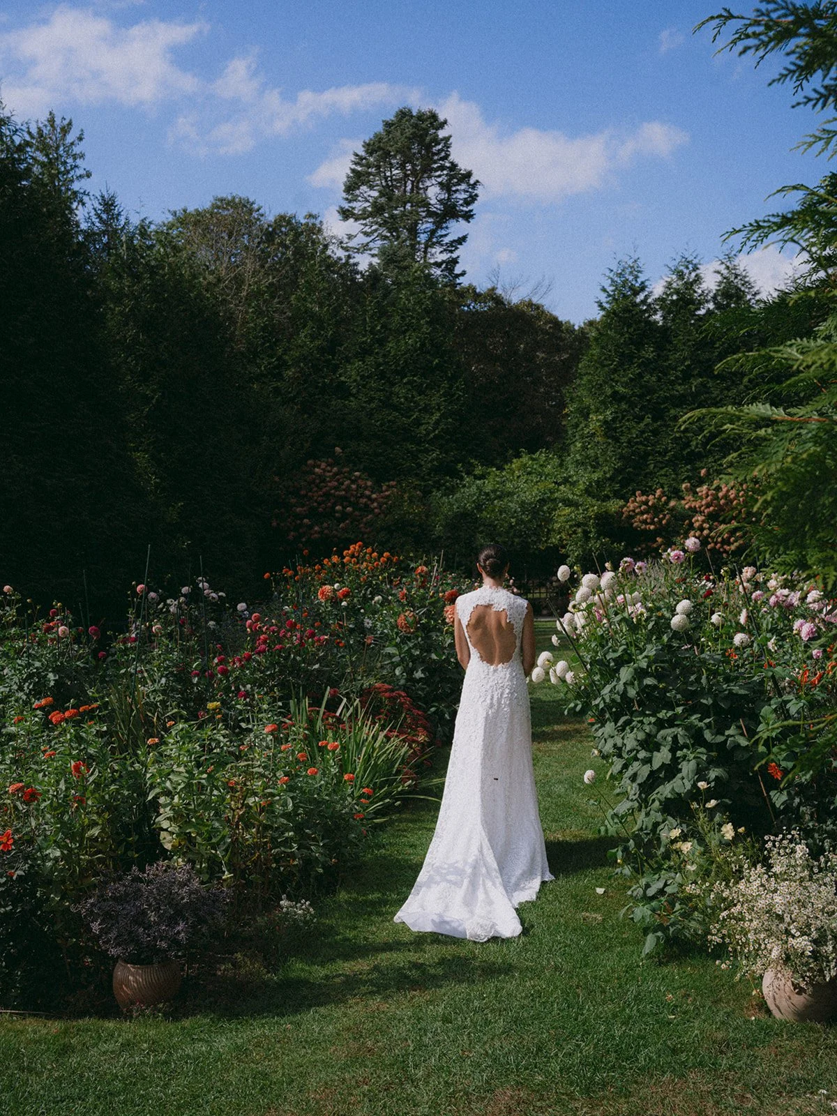 A woman in a white wedding dress walking through a lush garden with colorful flowers and tall trees under a partly cloudy sky during a wedding at Lambert's Cove Inn on Martha's Vineyard.