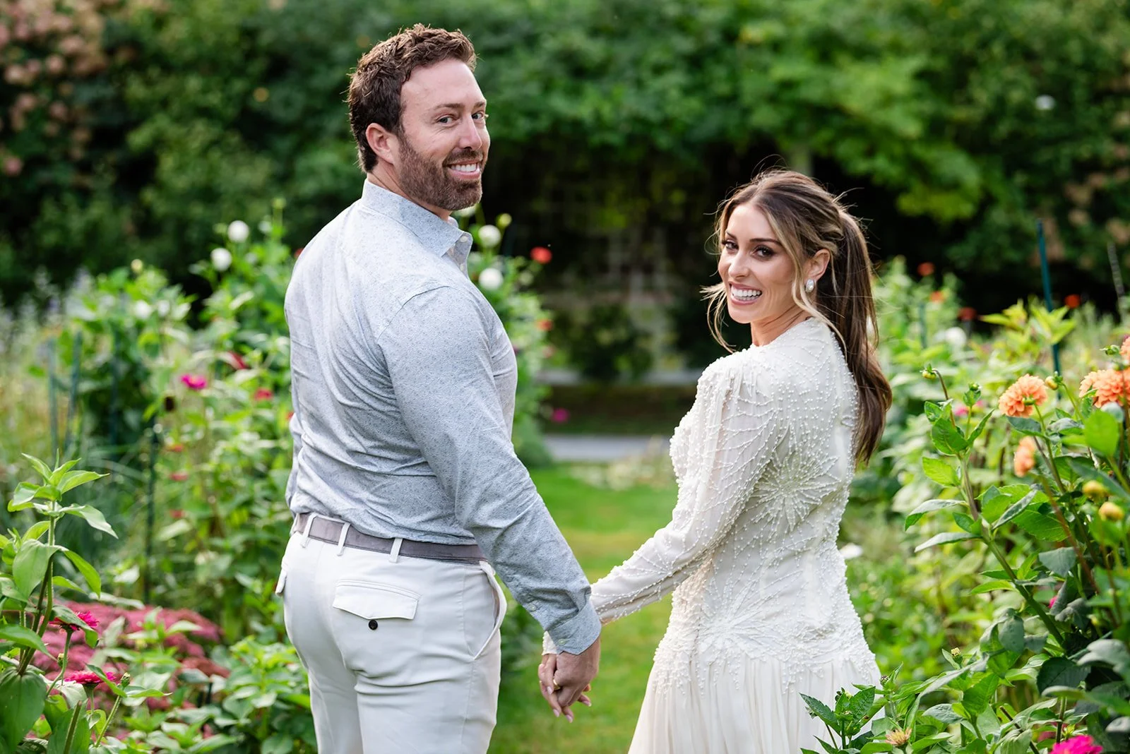 A smiling couple holding hands and looking back at the camera outside in a lush garden with blooming flowers at Lambert's Cove Inn on Martha's Vineyard.