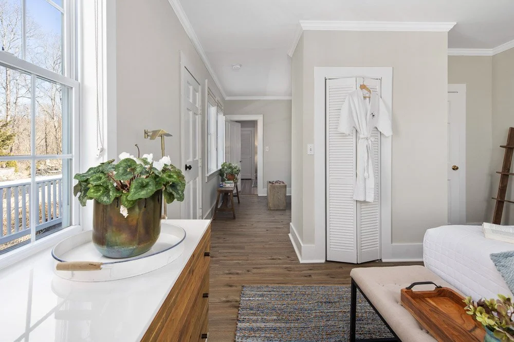 Interior view of a bright, minimalist bedroom with a large window, wooden flooring, a white bed, and a white robe hanging on a closet door.