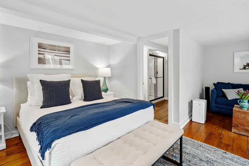 A bright bedroom with white walls, a large bed with navy blue pillows and a matching throw, a white nightstand with a lamp, a bench at the foot of the bed, and a sitting area with a blue sofa and potted plant. There is a framed photo on the wall and 