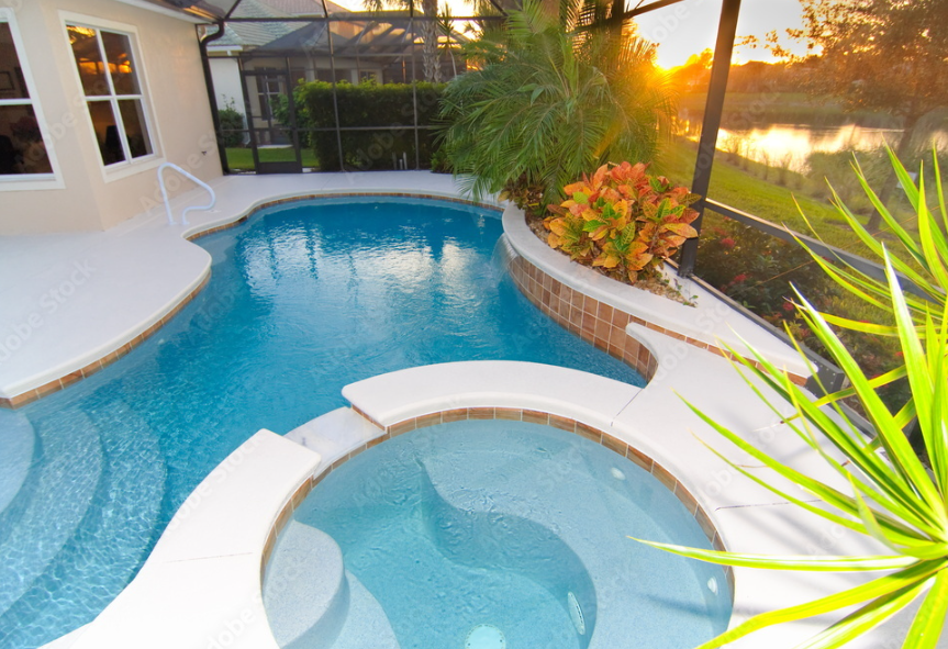 Swimming pool with hot tub, surrounded by tropical plants, during sunset, enclosed by a screened patio with view of a lake.