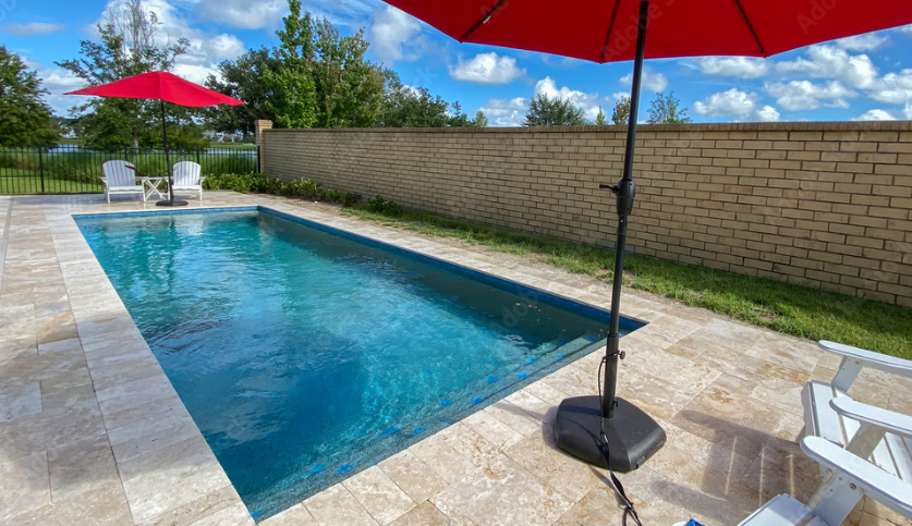 Small rectangular backyard swimming pool with chairs, umbrellas, and a brick wall on a sunny day.