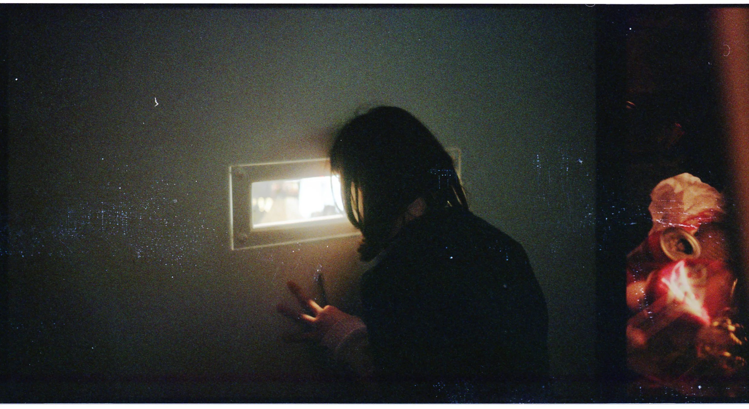 Person with dark hair looking at a small window in a dark room, with a faint reflection of a colorful object on the right side.