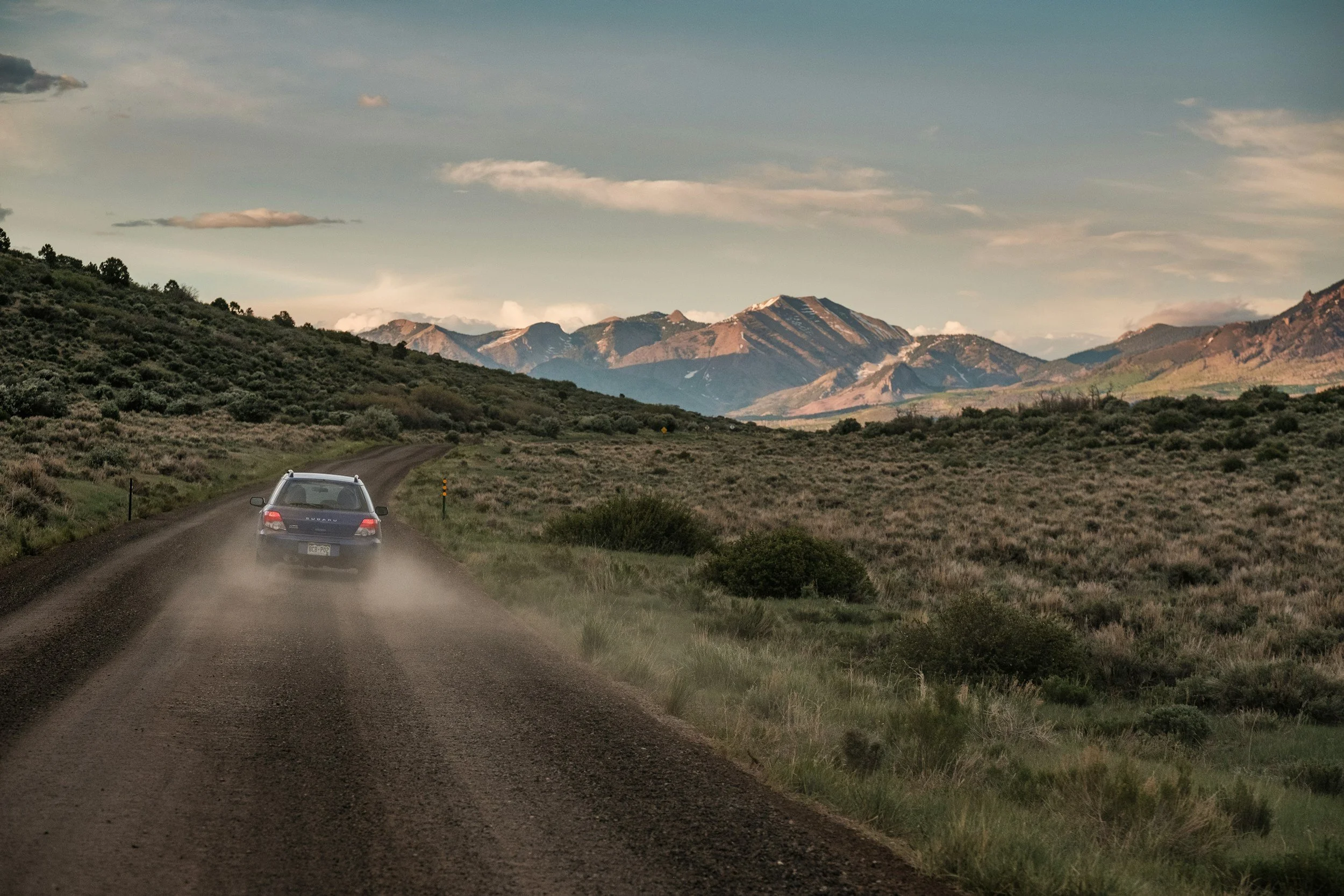 A car driving on a dirt road through a hilly landscape with mountains in the distance, under a partly cloudy sky.