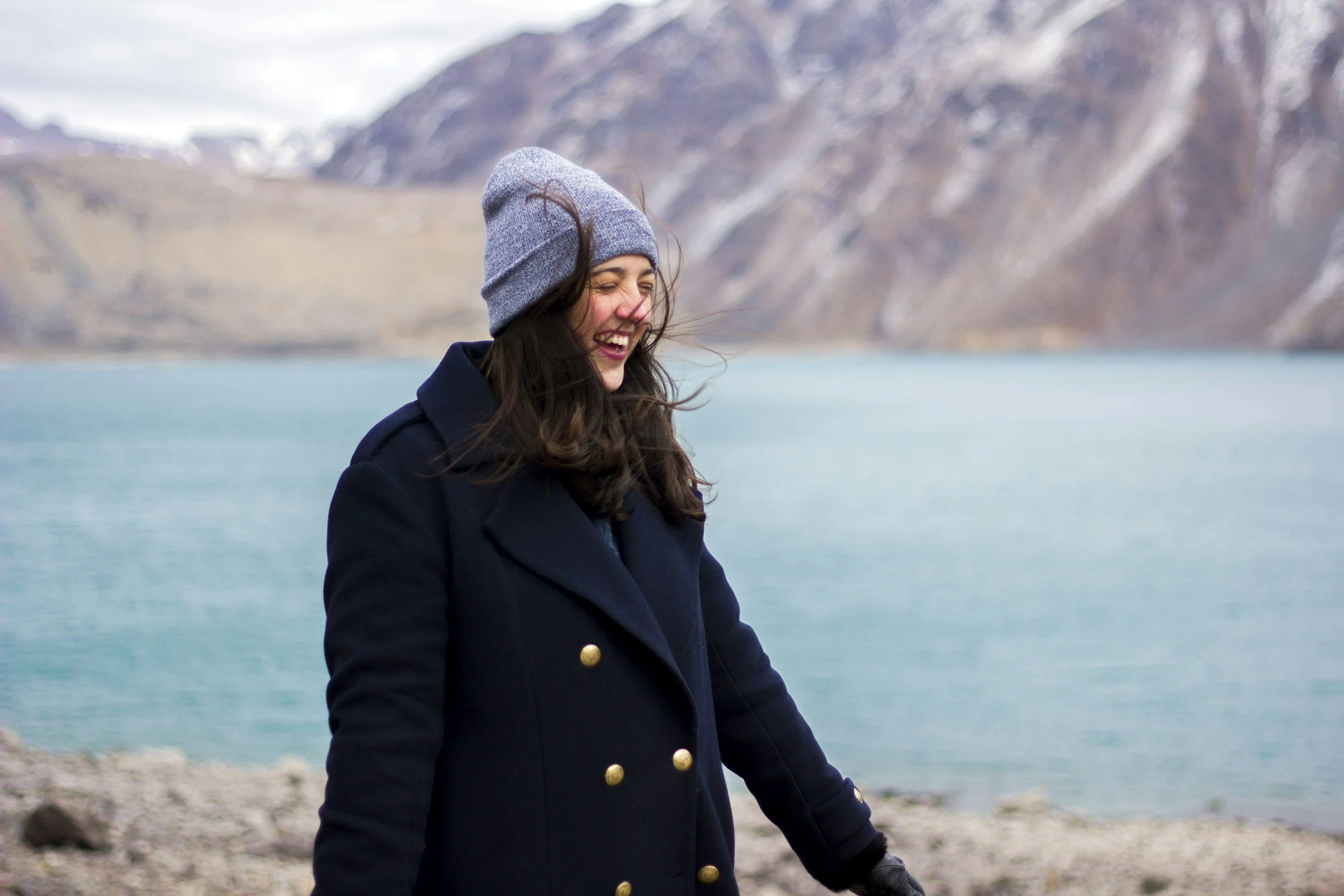 A woman with long brown hair, wearing a gray beanie and navy coat with gold buttons, standing near a body of water with mountains in the background, smiling and laughing.