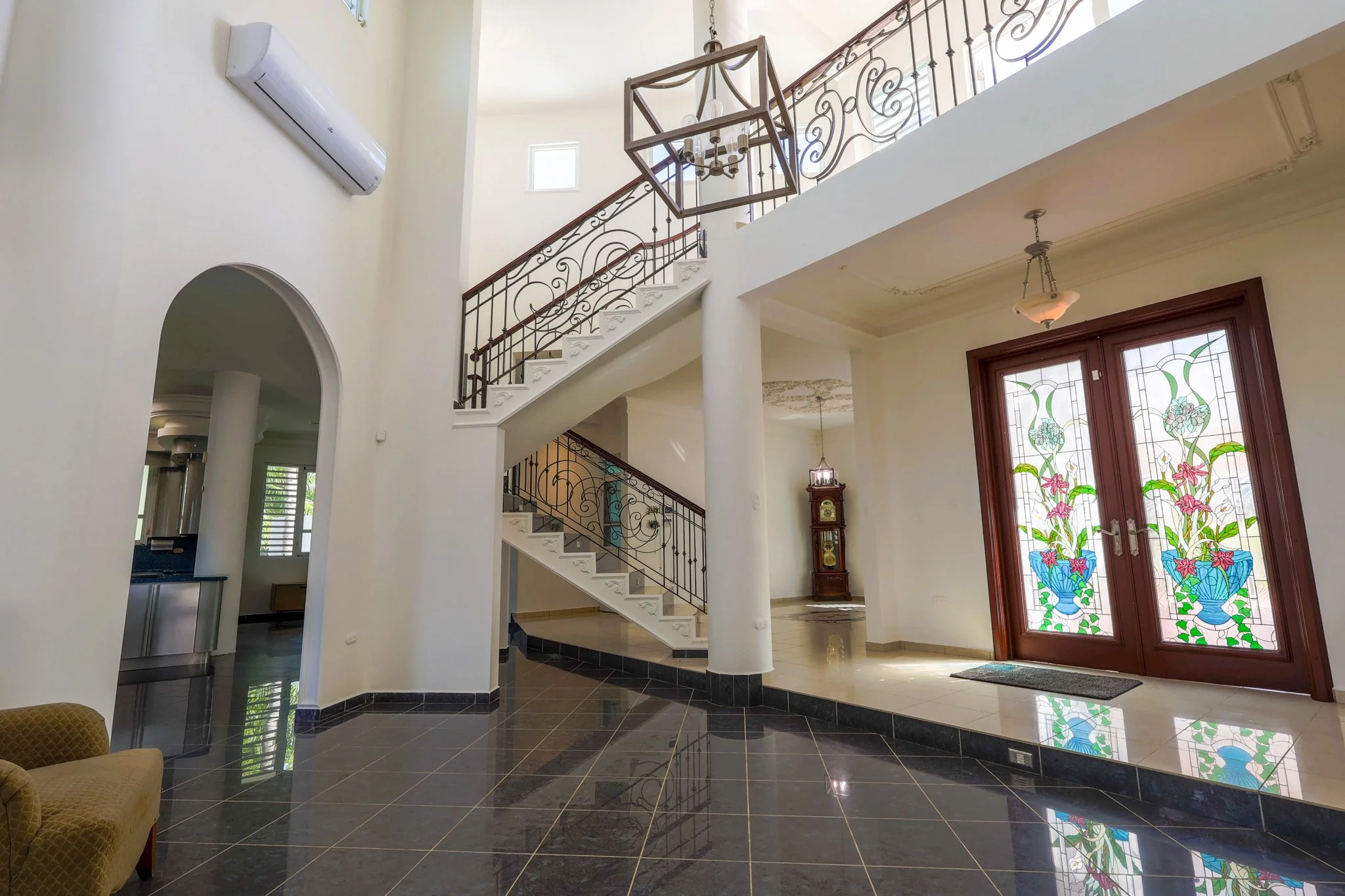 Interior view of a spacious house foyer with black tiled flooring, a staircase with decorative iron railings, stained glass doors with floral designs, and a grandfather clock.