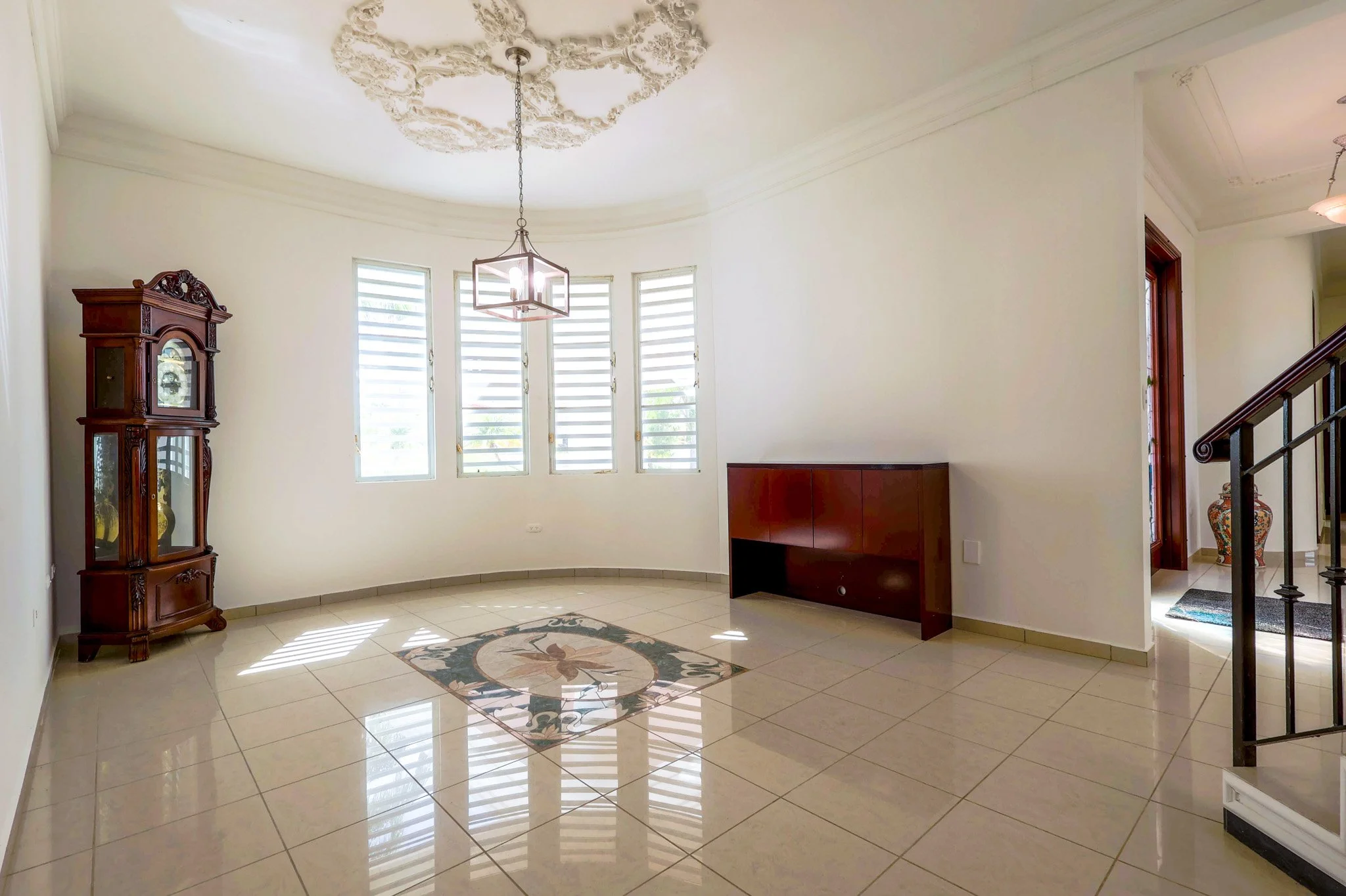 Empty living room with a chandelier, a wooden display cabinet, a red wooden cabinet, a decorative tile floor design, large windows with blinds, and a doorway with a vase visible in the hallway.