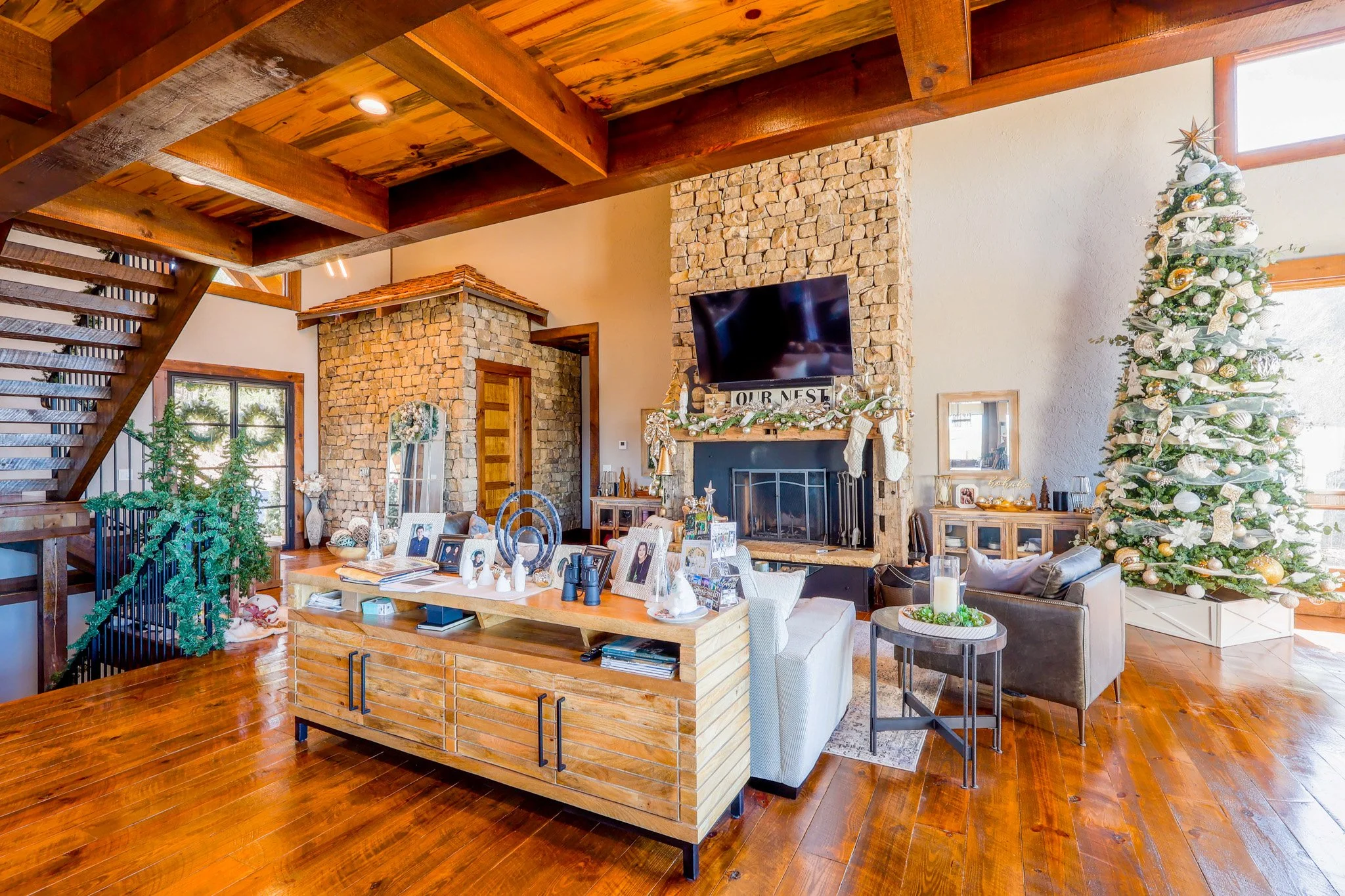 Festively decorated living room with a Christmas tree, a stone fireplace with stockings, a wooden staircase, and holiday decorations on a wooden table.