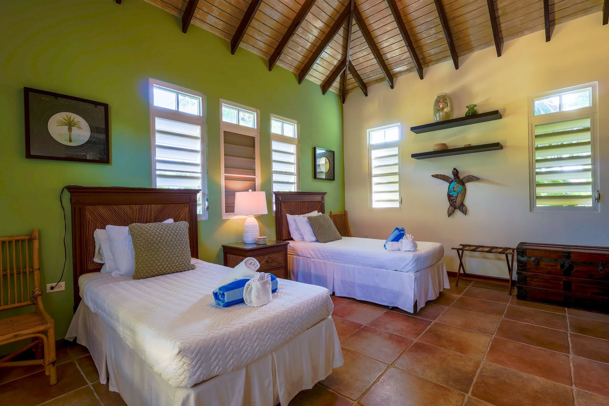 A bedroom with two single beds, each with white bedding and brown pillows, in front of green and white walls with multiple windows. There are wooden furniture pieces, a lamp, wall art, and decorative shelves.