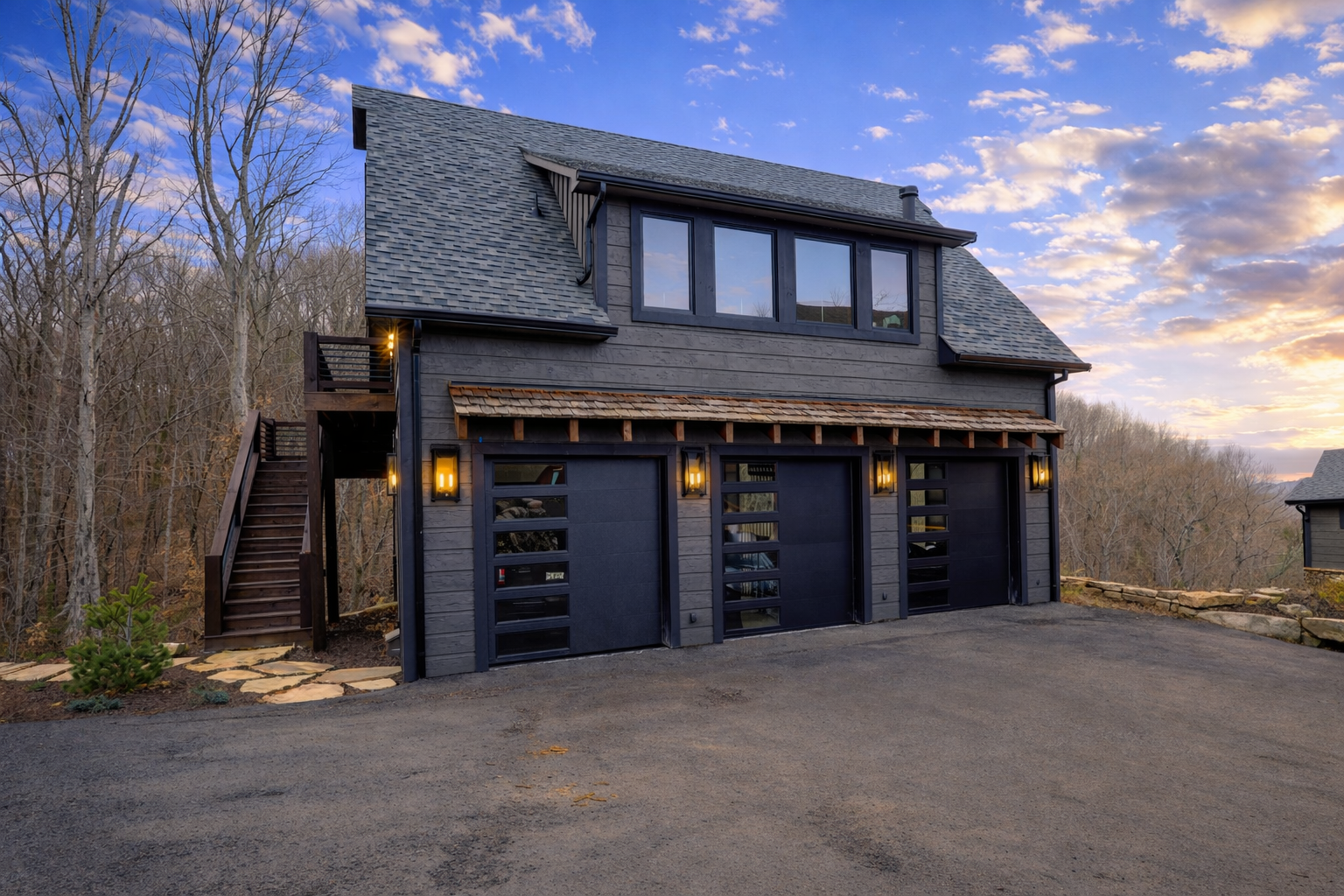 A modern two-story black house with three garage doors, exterior lights, and a staircase leading to the second floor, set against a backdrop of a sunset sky and barren trees.